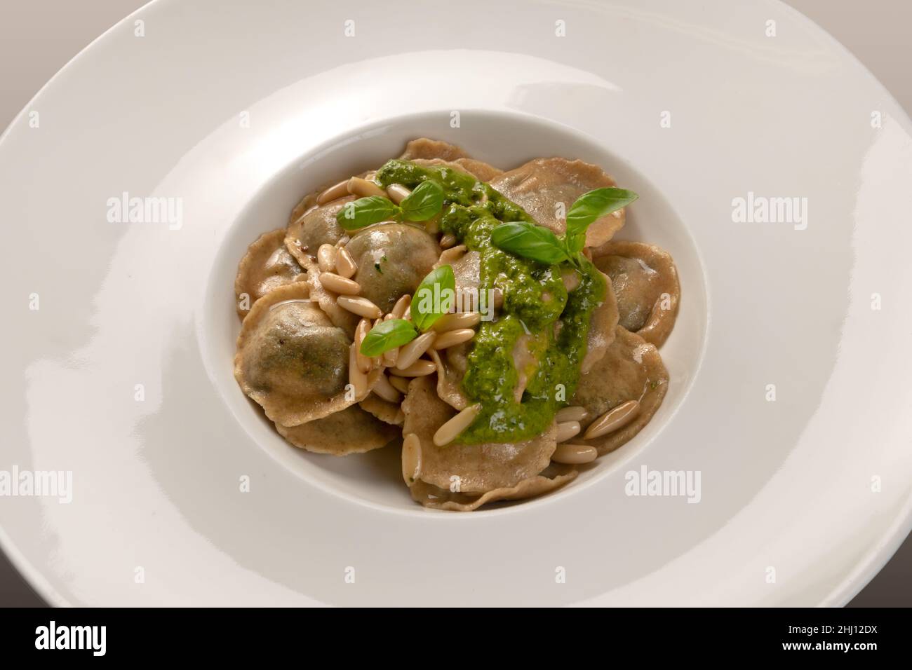 Raviolis ronds (tortelli) de farine complète avec pignons de pin et pesto génois et feuilles de basilic dans un plat blanc, gros plan Banque D'Images