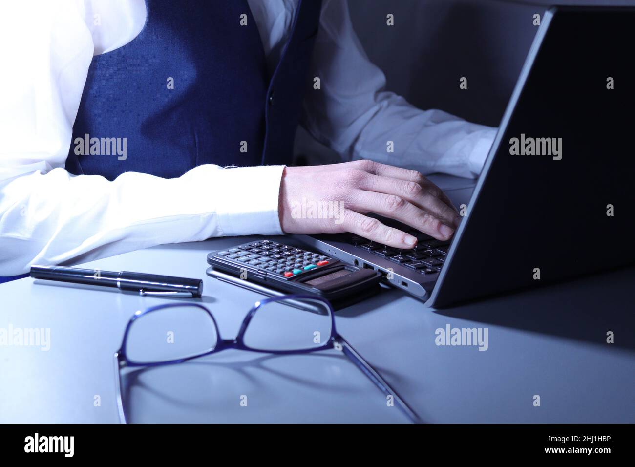 Employé de bureau en chemise blanche et gilet bleu travaillant à l'ordinateur portable avec des lunettes et une calculatrice sur le bureau Banque D'Images