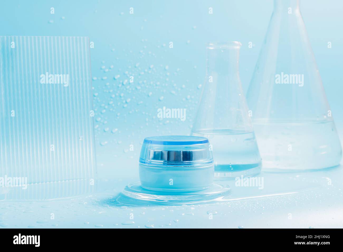 Crème cosmétique dans un pot, éclaboussures et gouttes d'eau sur fond bleu avec flacons en verre Banque D'Images