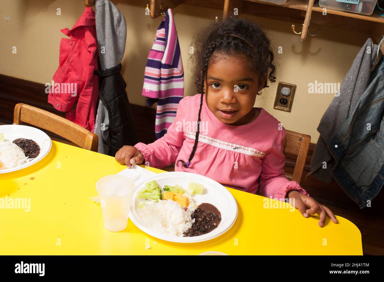 Éducation préscolaire jeune fille de 3-4 ans assise à table pour le déjeuner avec une assiette de nourriture devant ses haricots, riz, fromage, riz brocoli Banque D'Images