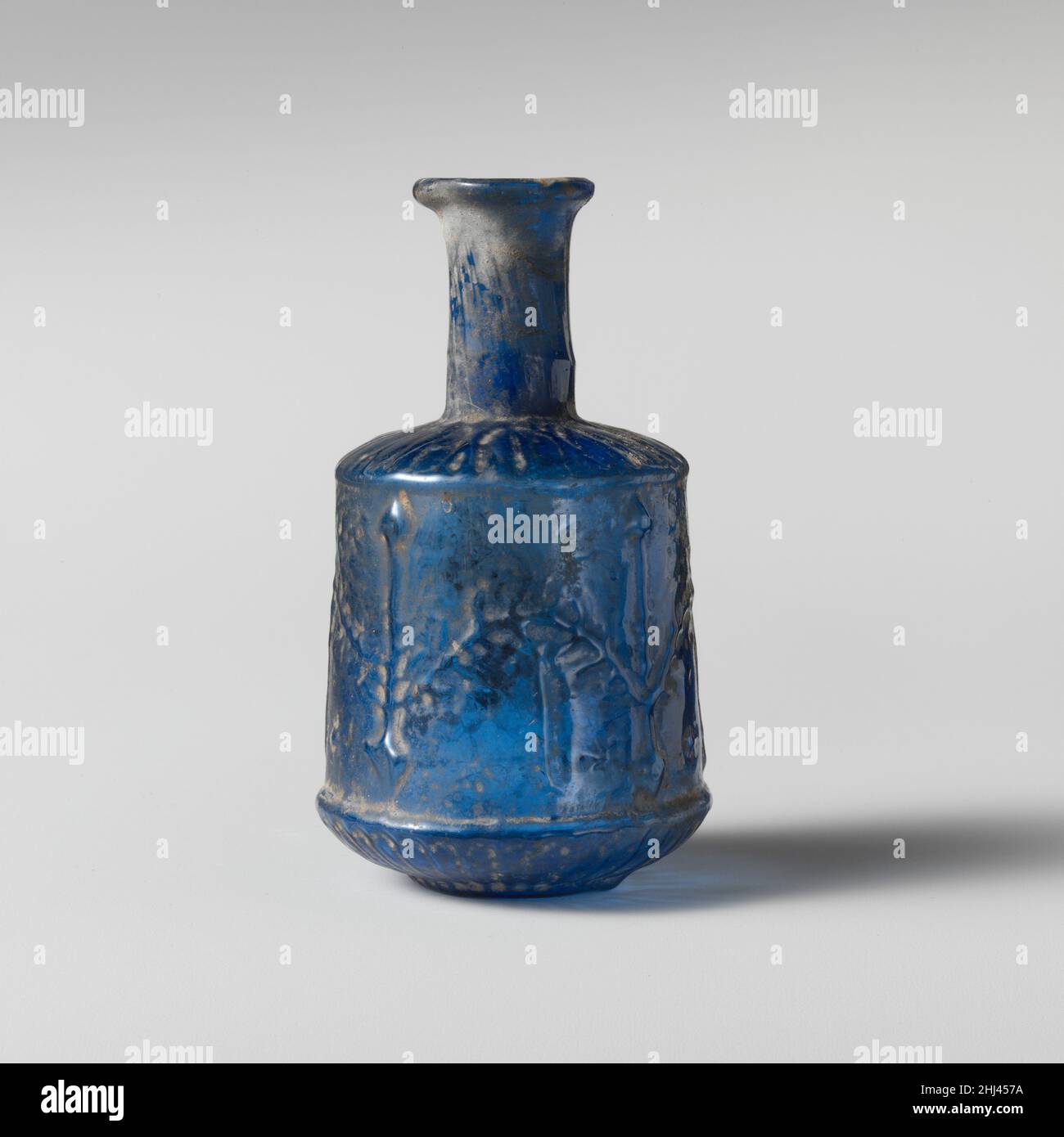 Flacon en verre 1st Century A.D. Roman translucide bleu cobalt.rebord éverté, partiellement replié ; col cylindrique, avec légère ligne horizontale de renflement vers la base ; épaule convexe inclinée avec arête extérieure proéminente ; corps cylindrique avec côtés légèrement extensibles vers le bas avec arête horizontale proéminente en dessous ; courbe convexe ;fond circulaire avec bossage central creux.Deux joints de moule s'exécutent de la partie inférieure du cou vers le bas, jusqu'au sommet de la proéminente arête horizontale ; une section en forme de cuvette distincte forme le corps inférieur et la base. Sur l'épaule, modèle non distinct de lignes radiantes ondulées ; sur le corps, suite Banque D'Images