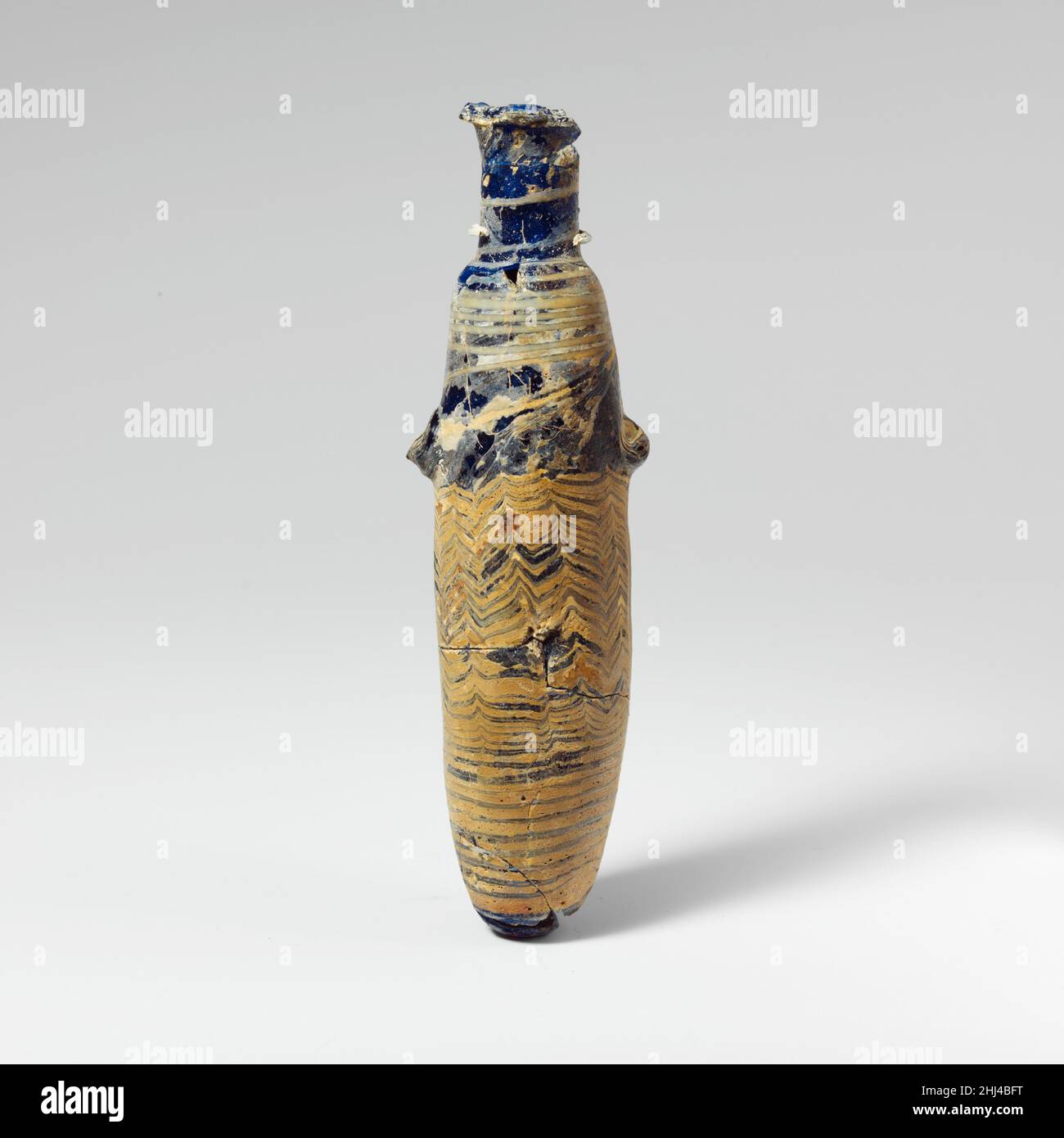Verre alabastron (flacon de parfum) mi-4th–début du 3rd siècle C.-B. grec, Méditerranée orientale ou italien Bleu cobalt translucide, avec poignées de la même couleur; pistes en blanc opaque (apparaissant bleu gris pâle) et jaune opaque.disque à rebord; col cylindrique haut; épaule inclinée; corps cylindrique légèrement bulbeux; petit fond convexe;Sur la carrosserie, deux grandes poignées à ergots, appliquées sur le motif de traînée, toutes deux avec des dents en dents de souris vers le haut.Une traînée blanche est appliquée sur le cou et s'enroule en spirale vers le bas du corps, se terminant par un cercle autour du fond; une autre jaune est appliquée sur le disque de jante et traîné sur le cou et Banque D'Images