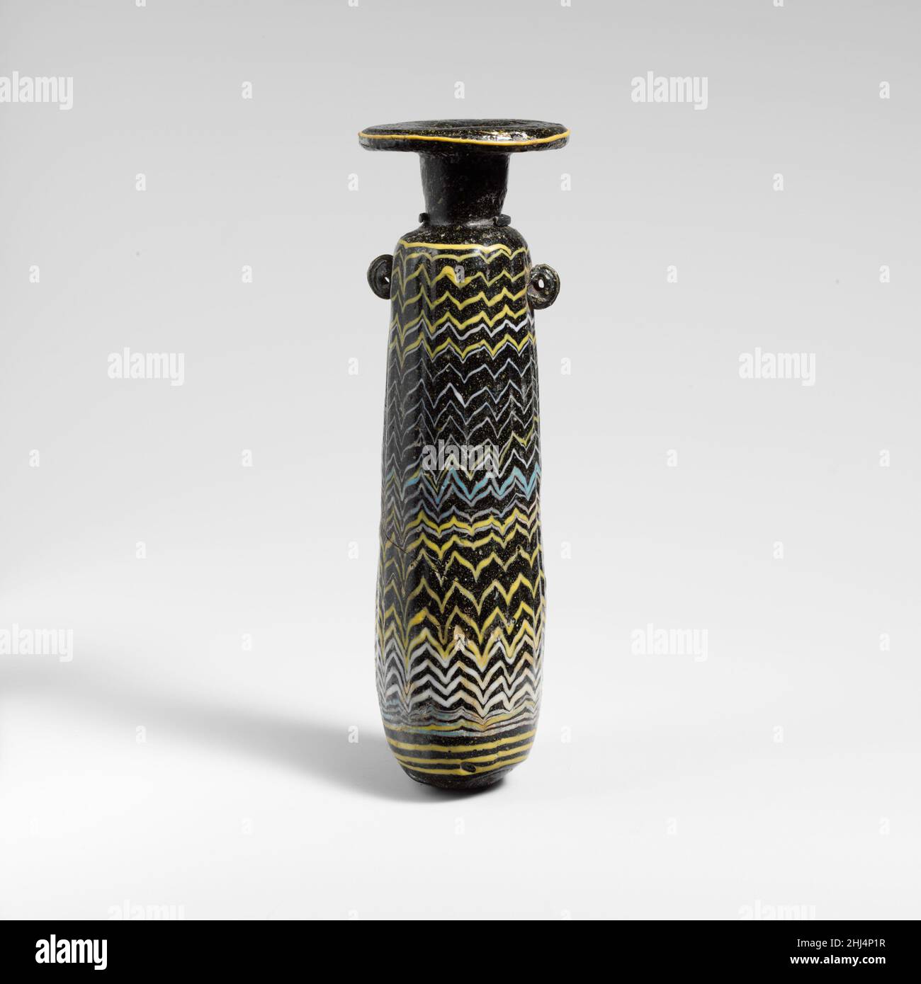 Verre alabastron (flacon de parfum) fin 4th–début 3rd siècle C.-B. Méditerranée orientale ou italien translucide vert très foncé (?), apparaissant noir opaque, avec poignées de la même couleur; traces en jaune opaque, blanc opaque et bleu turquoise opaque.disque à large rebord horizontal, avec bord arrondi épais; col cylindrique haut, effilée vers le bas; épaulement arrondi inégal;corps cylindrique à côtés droits,légèrement effilé vers le haut ; fond convexe inégal avec repères d'outillage et lissé dans un blob appliqué de verre de la même couleur ; sous l'épaulement, deux poignées verticales en anneau, l'une légèrement plus haute que l'autre, app Banque D'Images