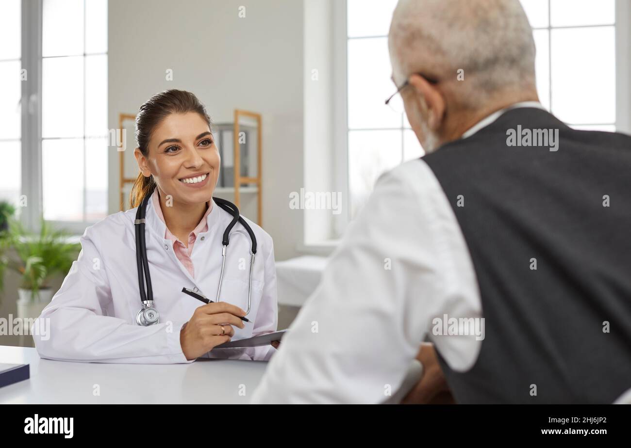 Une jeune femme sympathique médecin consulte un patient masculin senior et effectue des entrées dans sa carte médicale. Banque D'Images