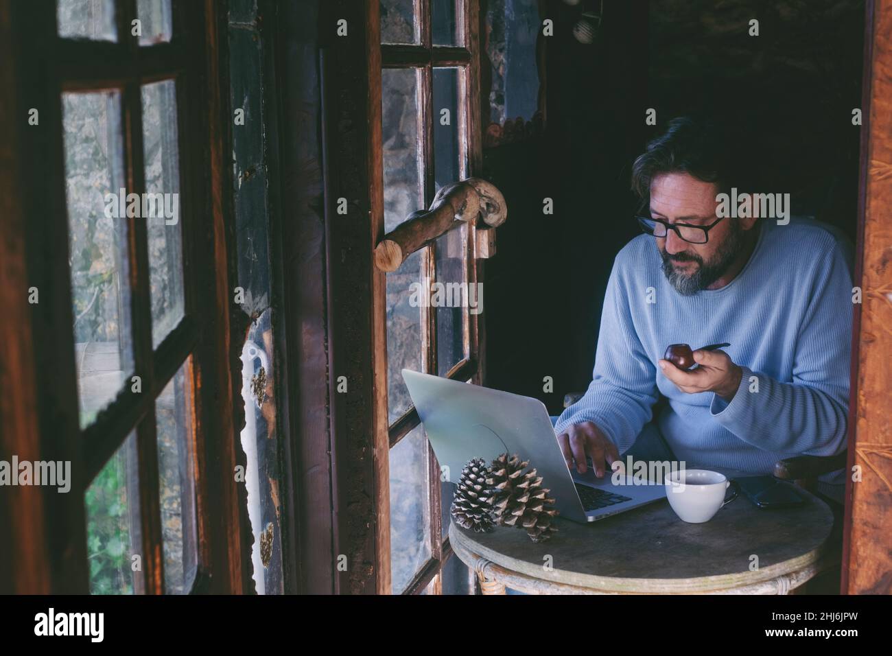 Homme mûr assis à la maison travaillant sur ordinateur portable et pipe fumeur.Alternative unique style de vie hommes personnes à l'intérieur dans chalet confortable en bois chalet.T Banque D'Images