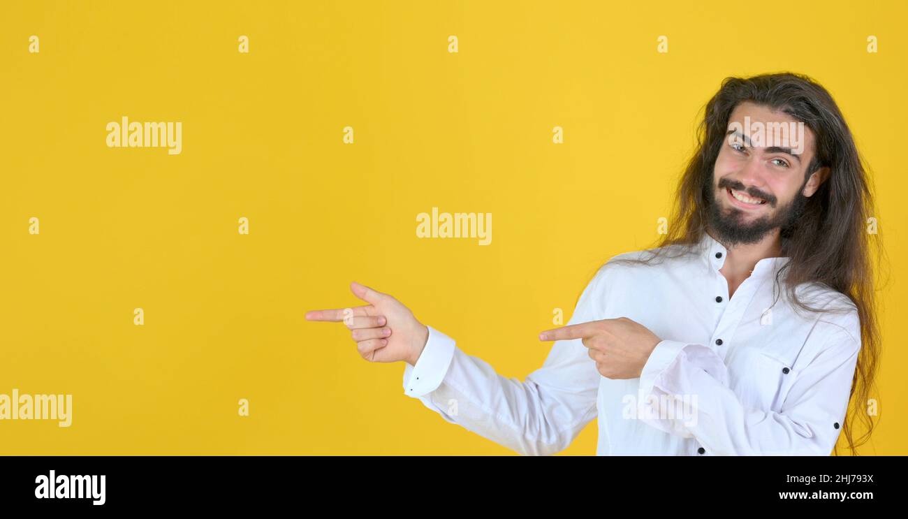 Jeune homme attrayant avec des cheveux longs et des sourires de barbe et pointant les doigts de côté à l'espace de copie pour votre texte isolé sur fond jaune studio Banque D'Images