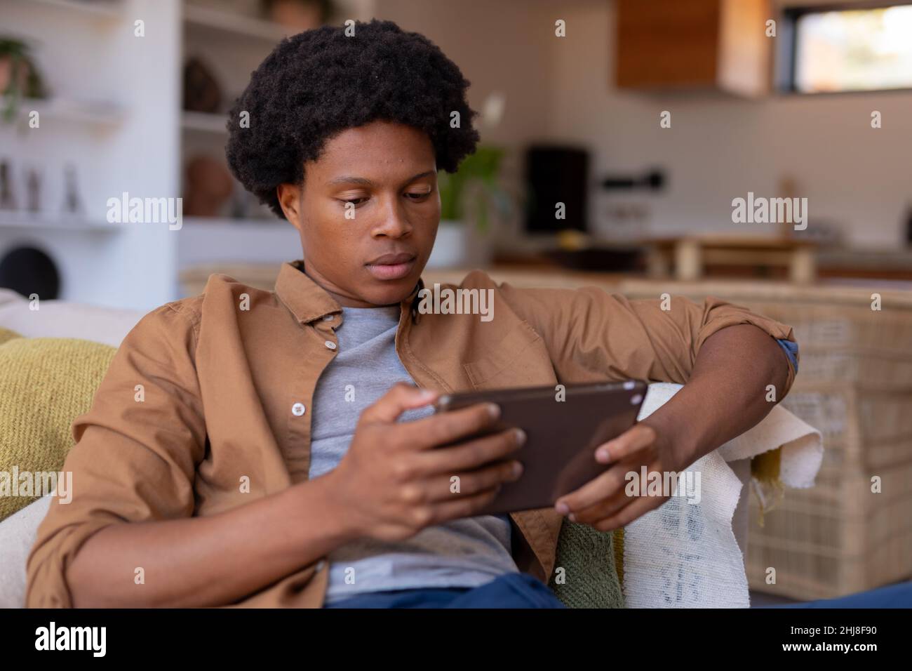 Jeune Afro-américain utilisant une tablette numérique tout en étant assis sur un canapé à la maison. Sans modification, style de vie, activité de loisirs, technologie sans fil, Concentrat Banque D'Images