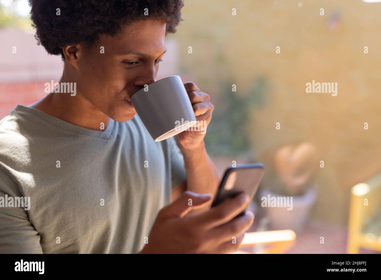 Gros plan sur un jeune homme afro-américain souriant qui boit du café tout en utilisant un smartphone à la maison. Sans modification, style de vie, loisirs, boisson, social Banque D'Images