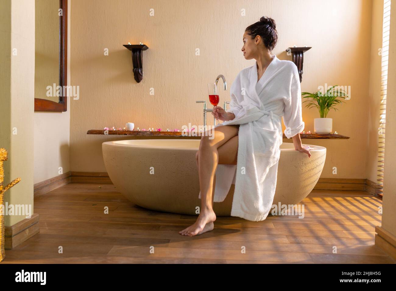 Jeune femme biraciale en peignoir avec vin rouge tout en étant assise sur la baignoire au spa. Spa, soins du corps, style de vie, vitiligo et bien-être. Banque D'Images