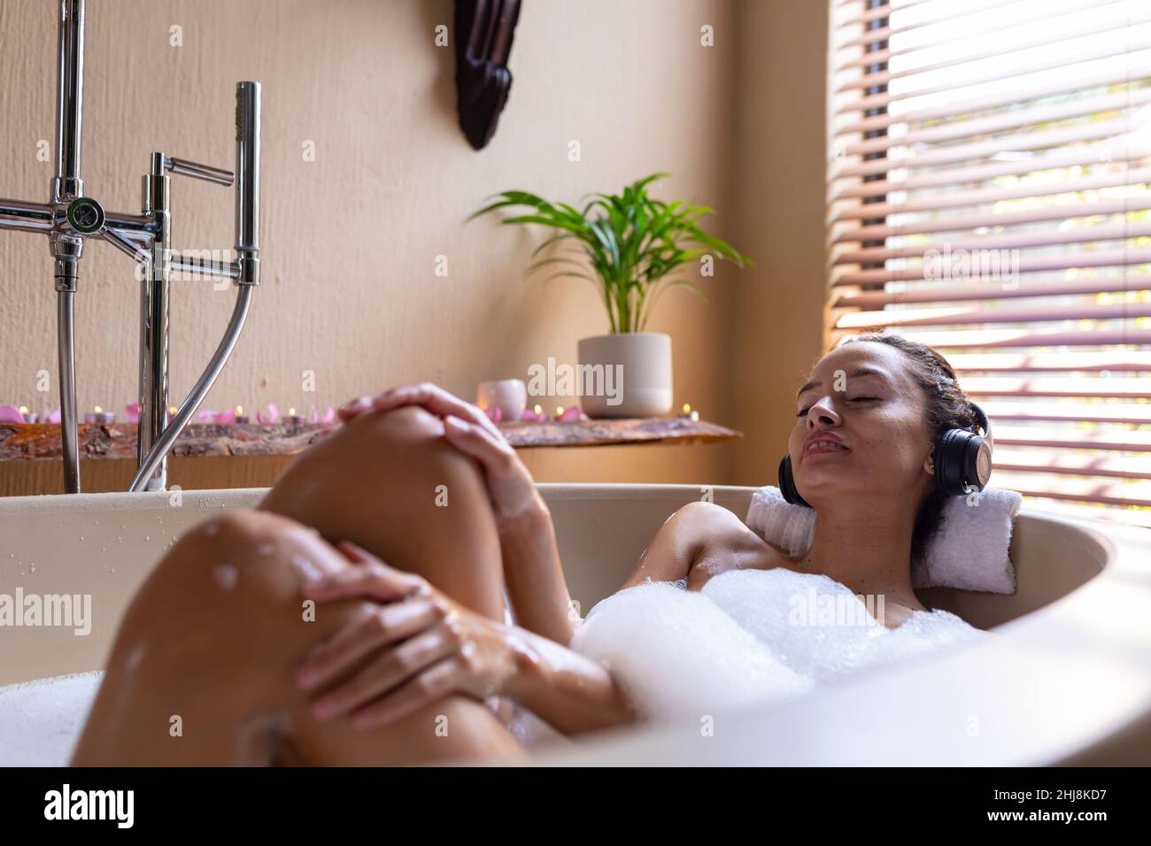 Jeune femme biracial écoutant de la musique à travers des écouteurs avec les yeux fermés dans la baignoire savonnante au spa. Unaltered, spa, soins du corps, sensualité, style de vie,vi Banque D'Images