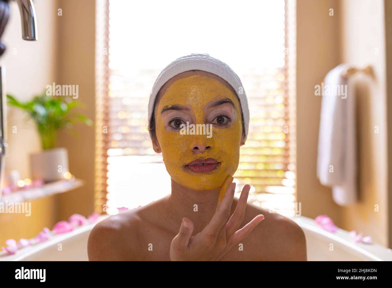 Gros plan portrait de la jeune femme biraciale appliquant un masque facial jaune dans la baignoire au spa. Non modifié, spa, soins du corps, style de vie, vitiligo et bien-être. Banque D'Images