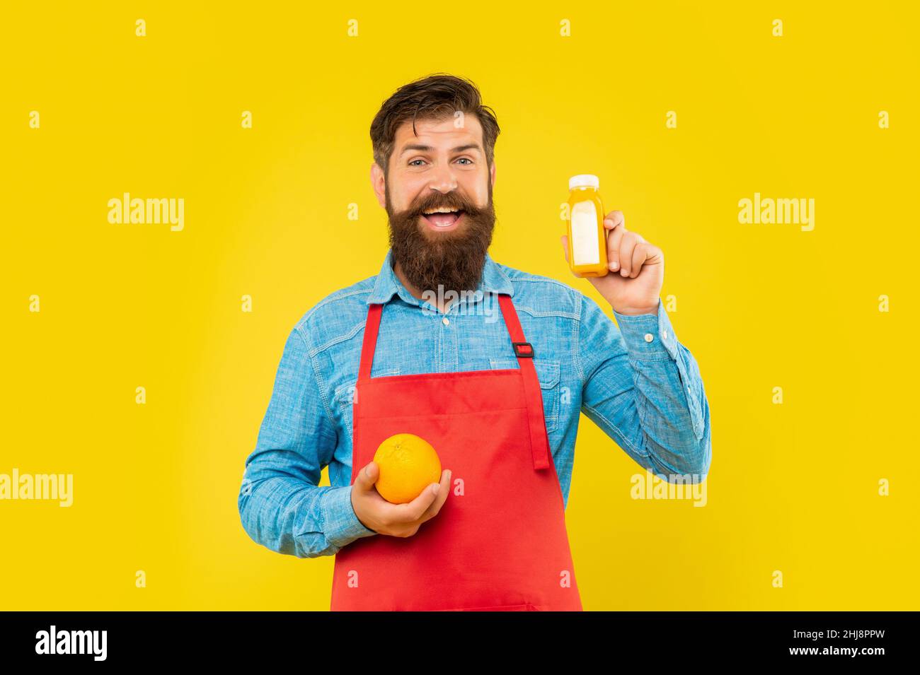 Homme heureux en tablier tenant orange et bouteille de jus fond jaune, barman Banque D'Images