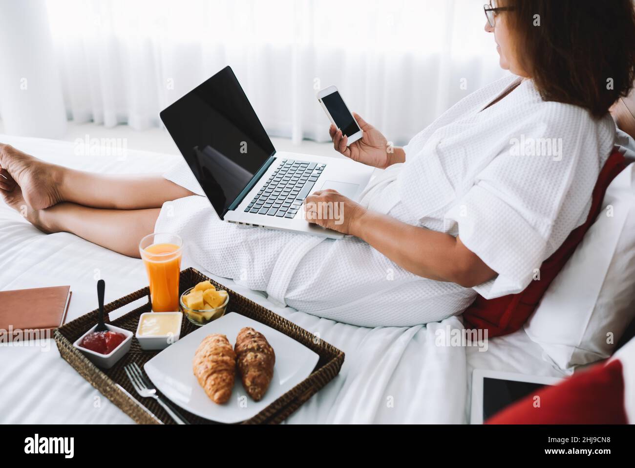 Rognez la femme en utilisant des gadgets sur le lit de l'hôtel avec le petit déjeuner Banque D'Images