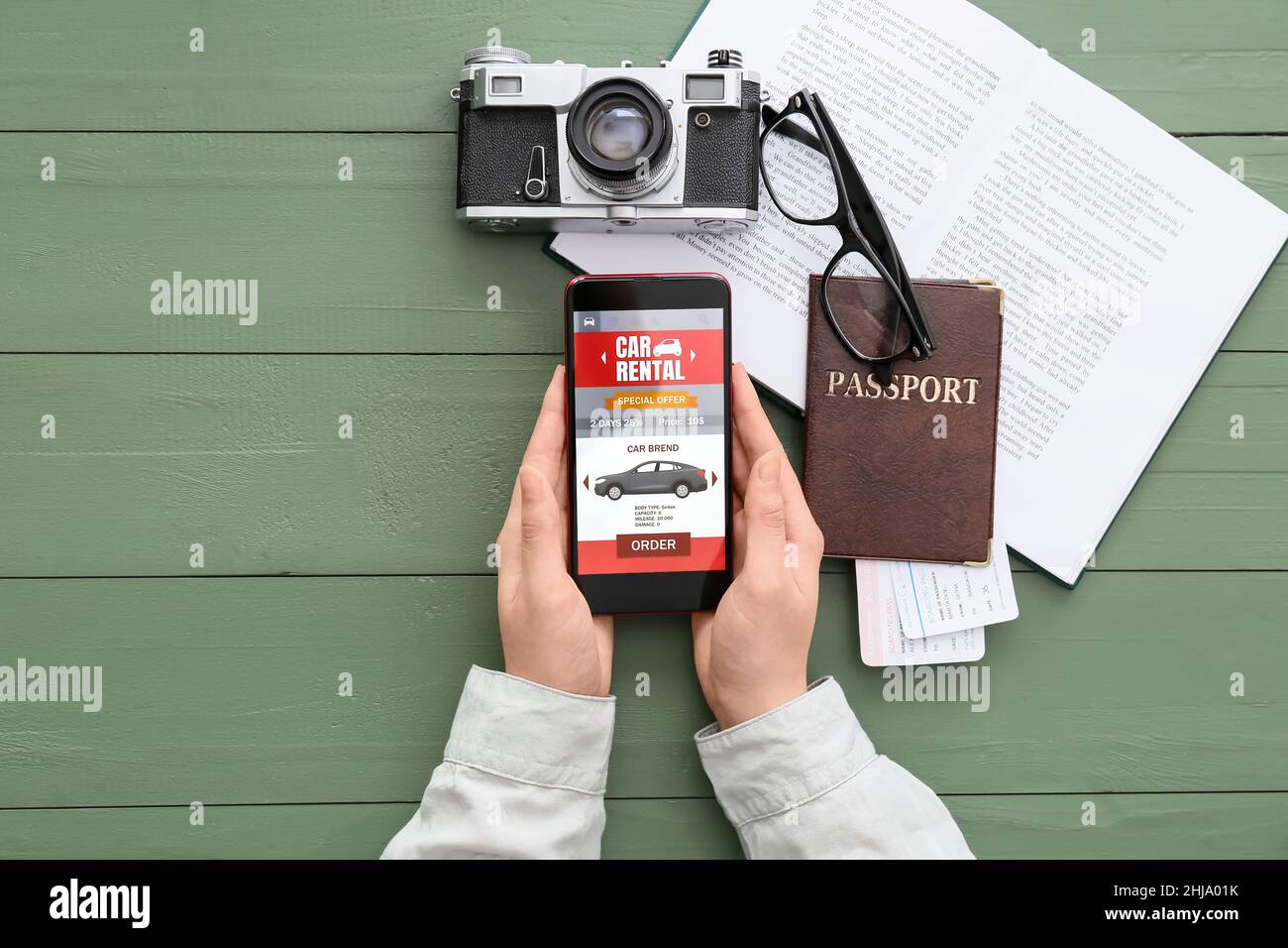 Femme tenant un téléphone portable avec une application de location de voiture ouverte, un appareil photo, un passeport et un livre sur fond de bois vert Banque D'Images