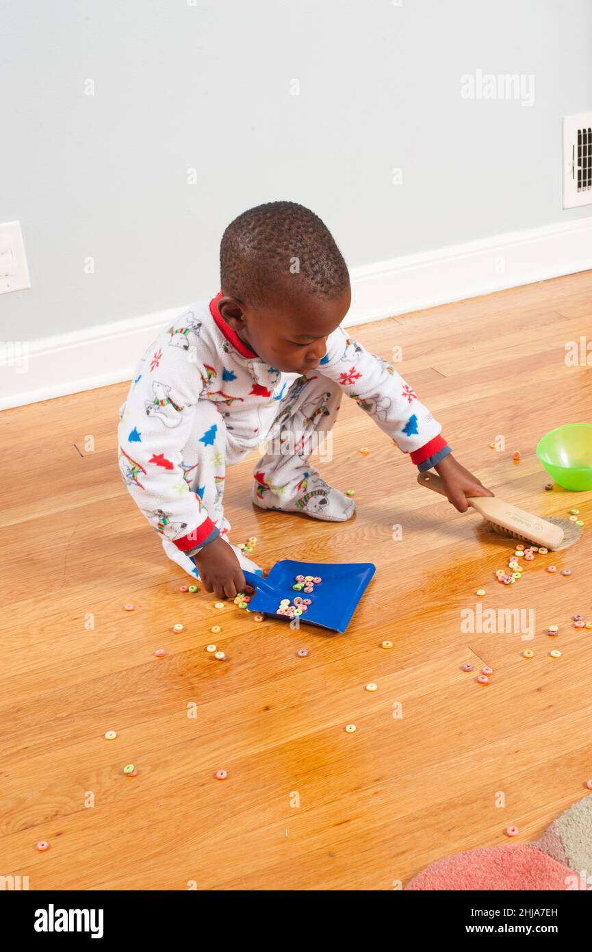 Un tout-petit garçon à la maison utilise un bac à poussière et une brosse pour nettoyer les céréales déversées sur le sol, portant un pyjama Banque D'Images