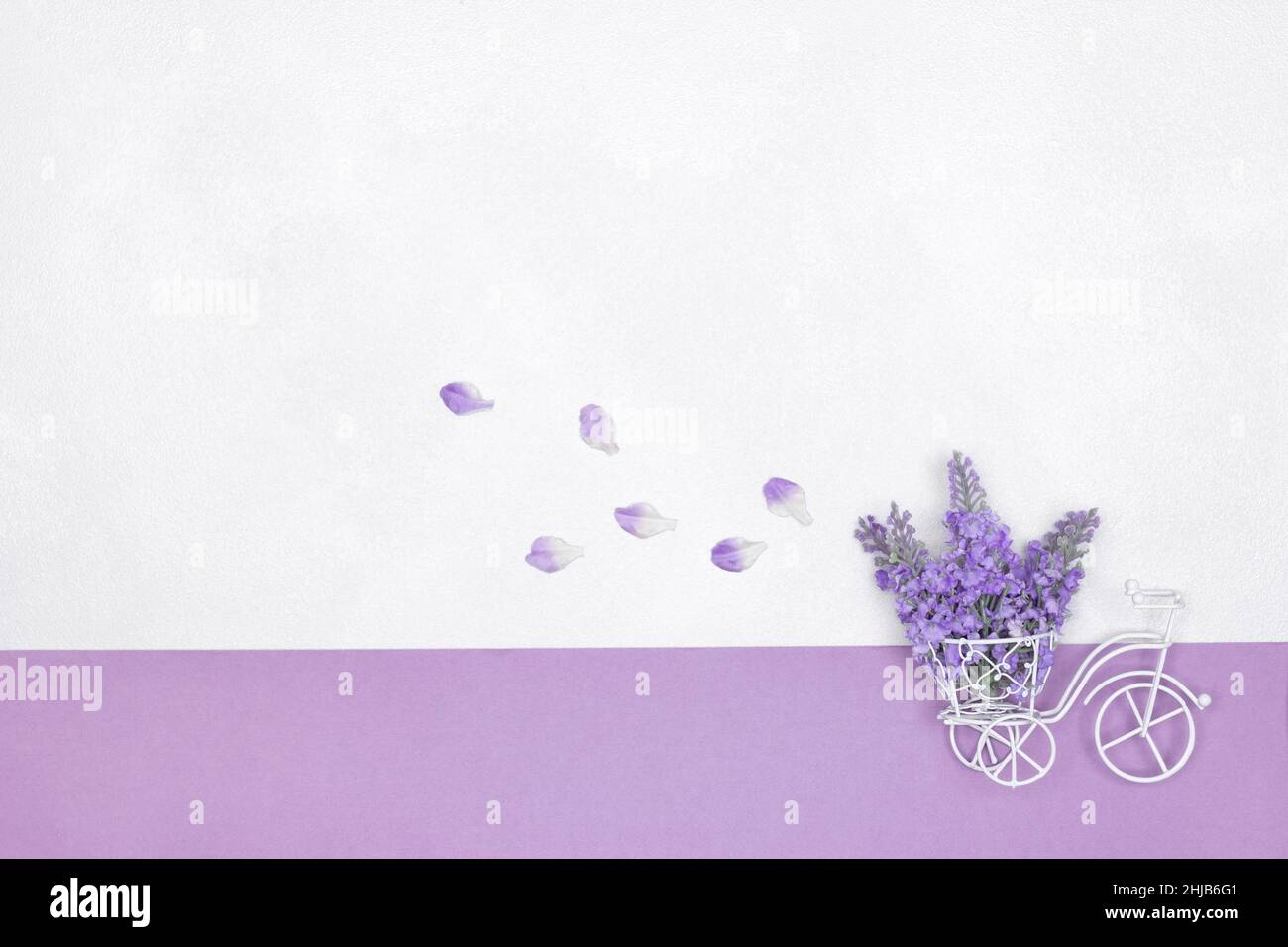 Petit vélo blanc jouet avec bouquet de fleurs violettes dans le panier et pétales volants derrière.Arrière-plan de printemps mignon.Copier l'espace Banque D'Images