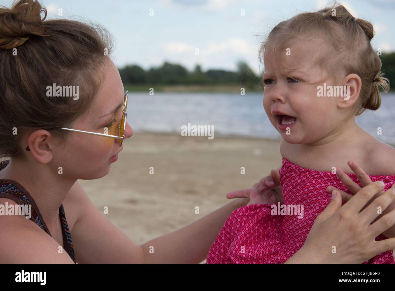 Enfant caucasien capricieux de deux ans qui est calmé par une jeune femme sur la plage d'été Banque D'Images