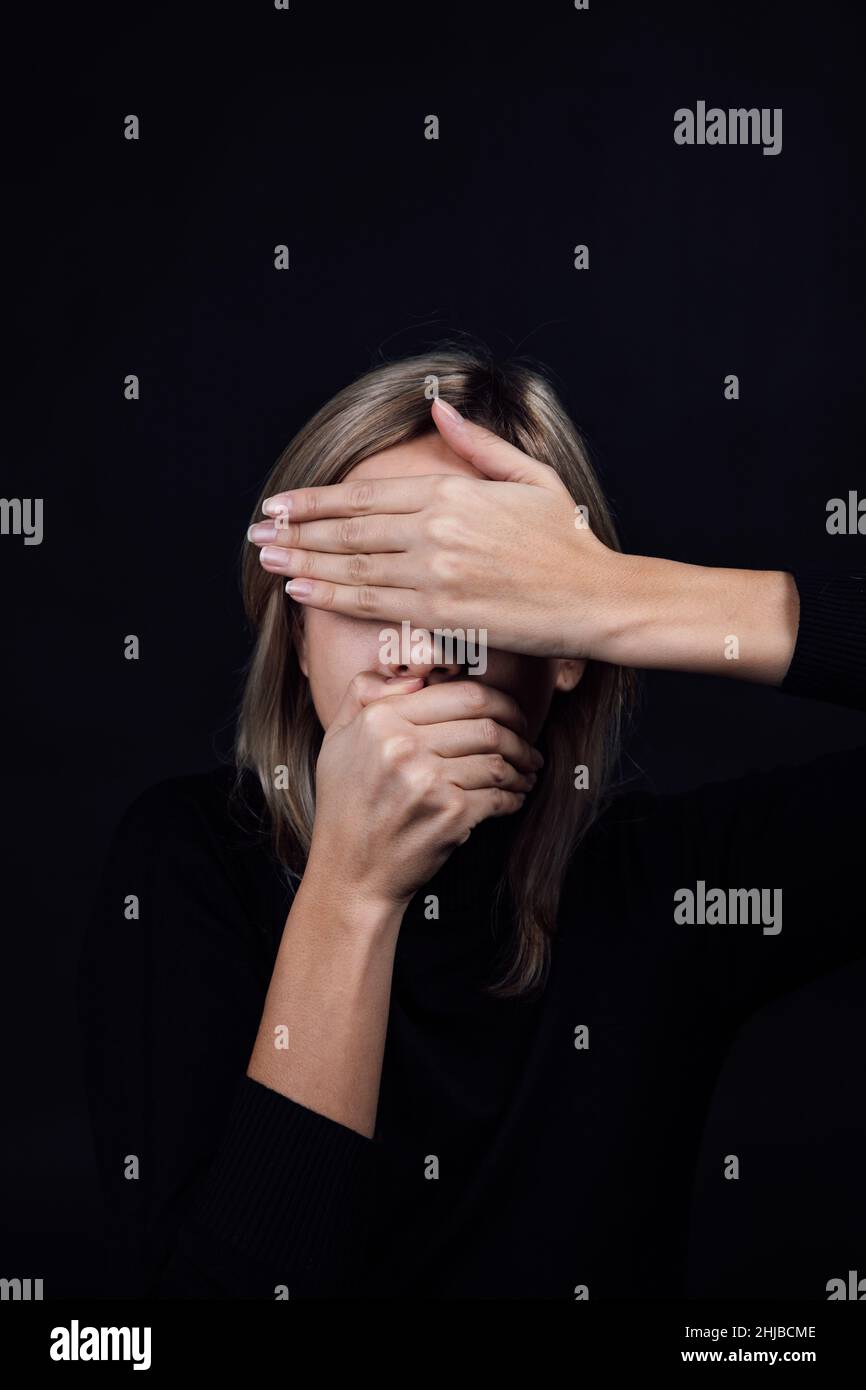 Femme opprimée aux mains cachant les yeux et la bouche portant un chemisier noir sur fond noir.Victime d'abus physique et psychologique.Éclairage au gaz Banque D'Images