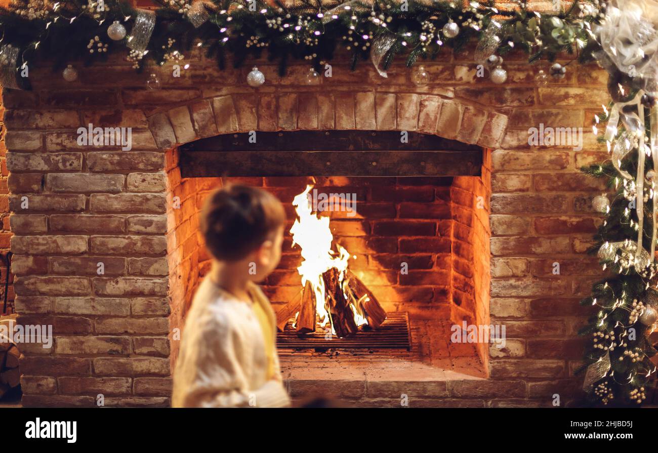 Petit garçon près d'une cheminée en brique décorée d'une guirlande de vacances, foyer sélectif.Enfant assis au feu dans une maison de campagne confortable pendant les vacances d'hiver, r Banque D'Images
