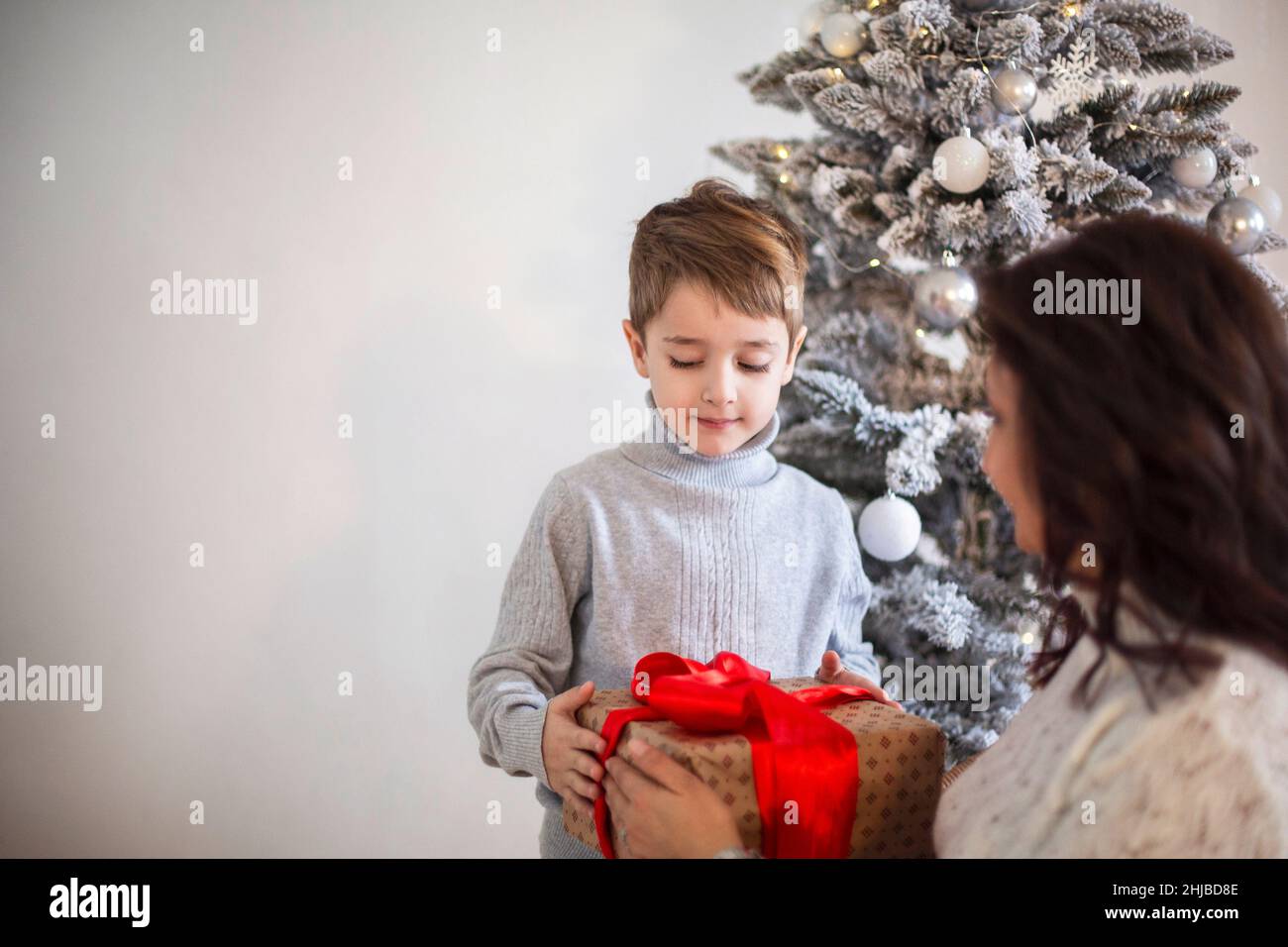 Mignon petit garçon recevant le cadeau de Noël de la grand-mère tout en se  tenant près de l'arbre artificiel décoré à la maison.J'adore donner un cadeau  de Noël emballé Photo Stock -