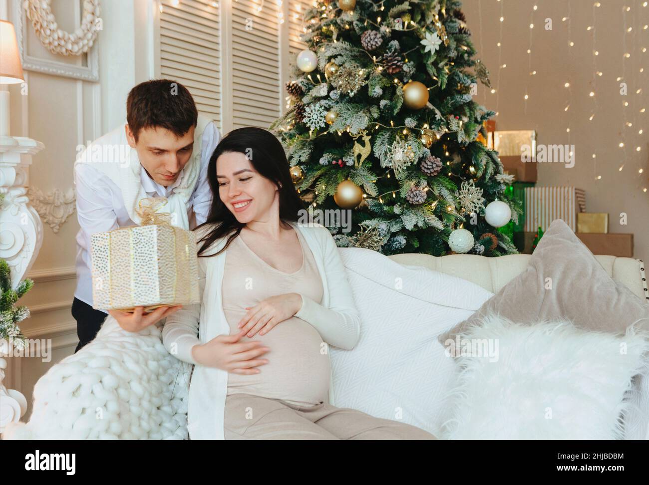 Jeune mari aimant donnant cadeau de vacances à une femme enceinte heureuse surprise tout en célébrant Noël ou nouvel an ensemble.Future jeune famille par Banque D'Images
