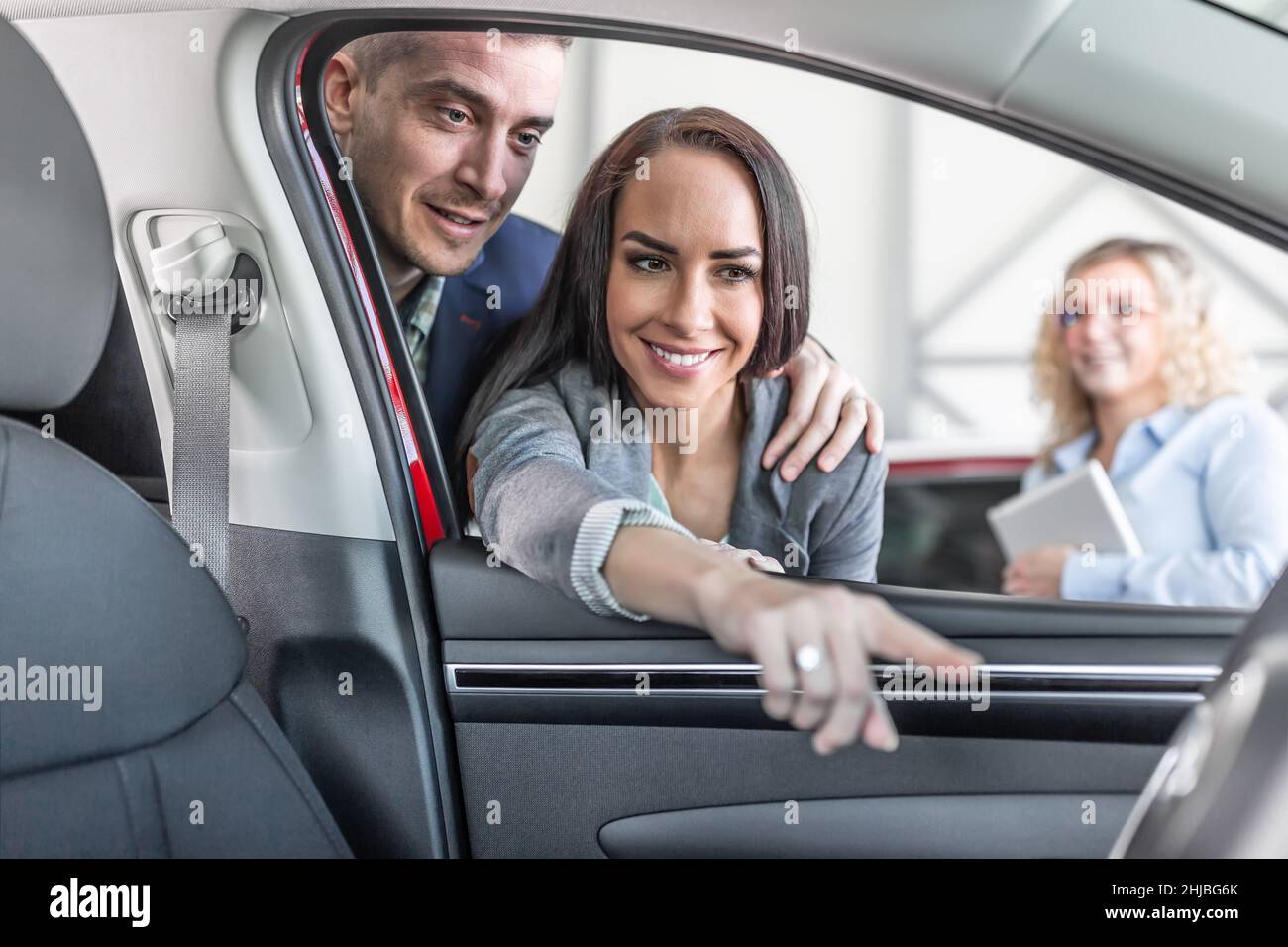 Un jeune couple vérifie les caractéristiques de l'intérieur d'une nouvelle voiture tandis que le directeur des ventes se tient de côté dans la concession. Banque D'Images