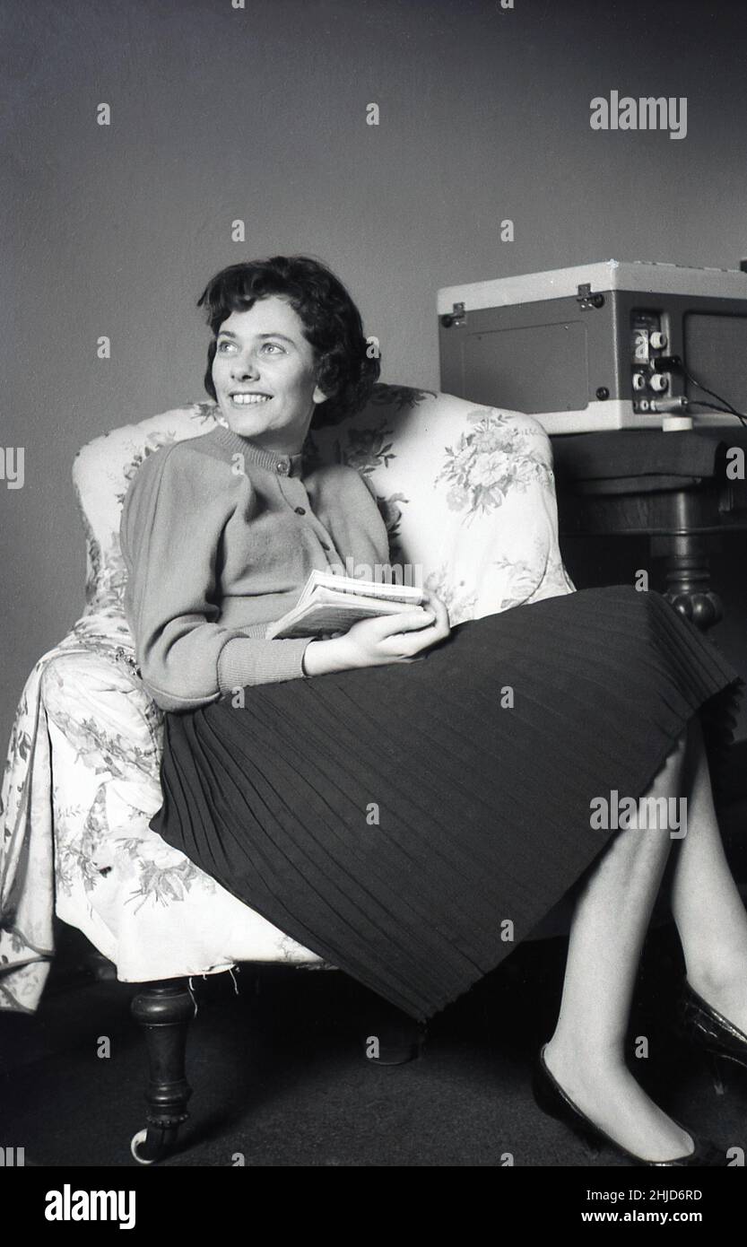 1950s, historique, à l'intérieur d'une pièce, assis dans une chaise, une jeune femme élégamment habillée portant un chemisier en laine et une jupe plissée, avec un livre de poche sur les genoux, souriant pour sa photo.Un lecteur de disque ou de gramophone portable est sur une table à côté d'elle, Angleterre, Royaume-Uni Banque D'Images