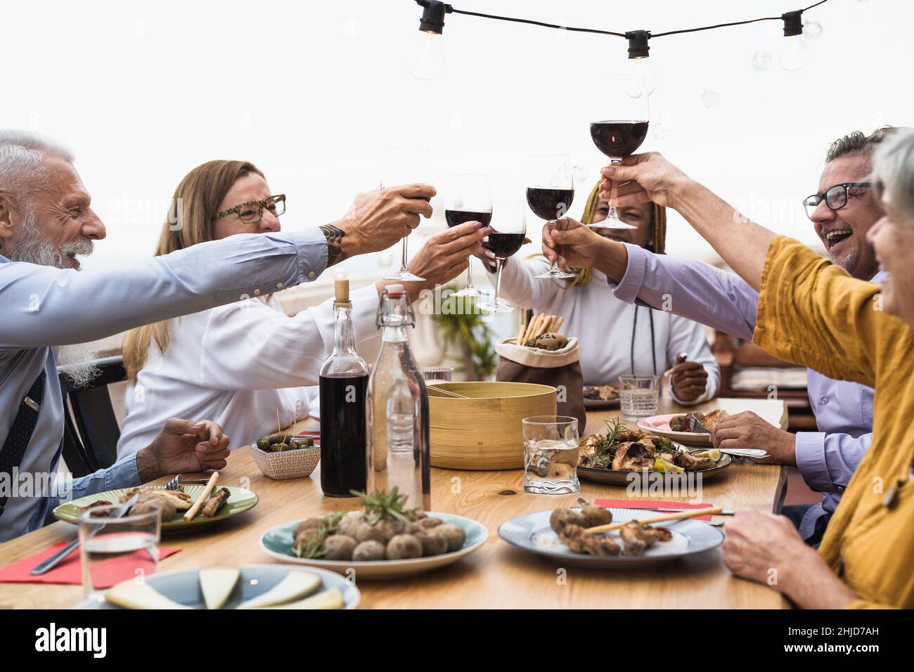 Des amis seniors multiraciaux s'amusent à manger ensemble et à faire du toaster avec Dîner avec vin rouge sur le patio de la maison - repas et vacances concept Banque D'Images