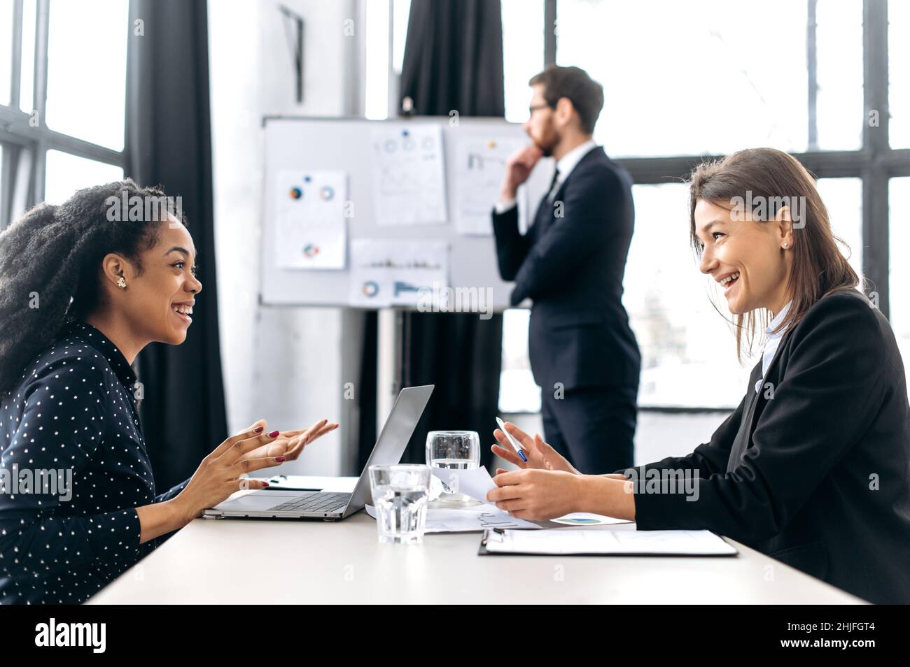 Un groupe de collègues multiraciaux travaille dans un bureau moderne.Deux femmes réussies sont en négociation, discuter de projet d'affaires, la planification de la stratégie financière, sourire, l'homme se tient sur le fond Banque D'Images