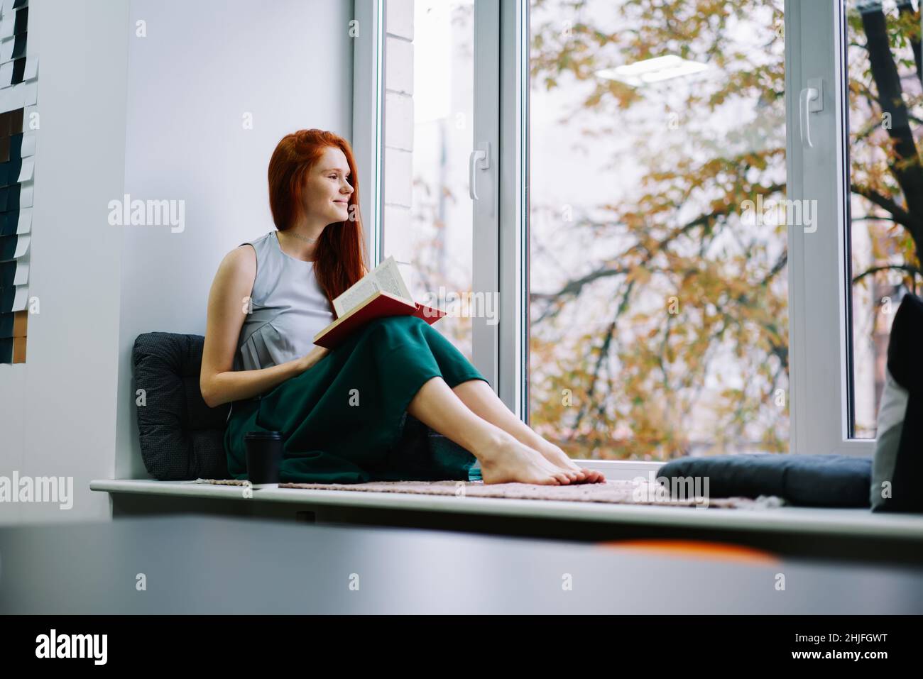 Femme rêveuse assise sur le rebord de la fenêtre avec un livre Banque D'Images