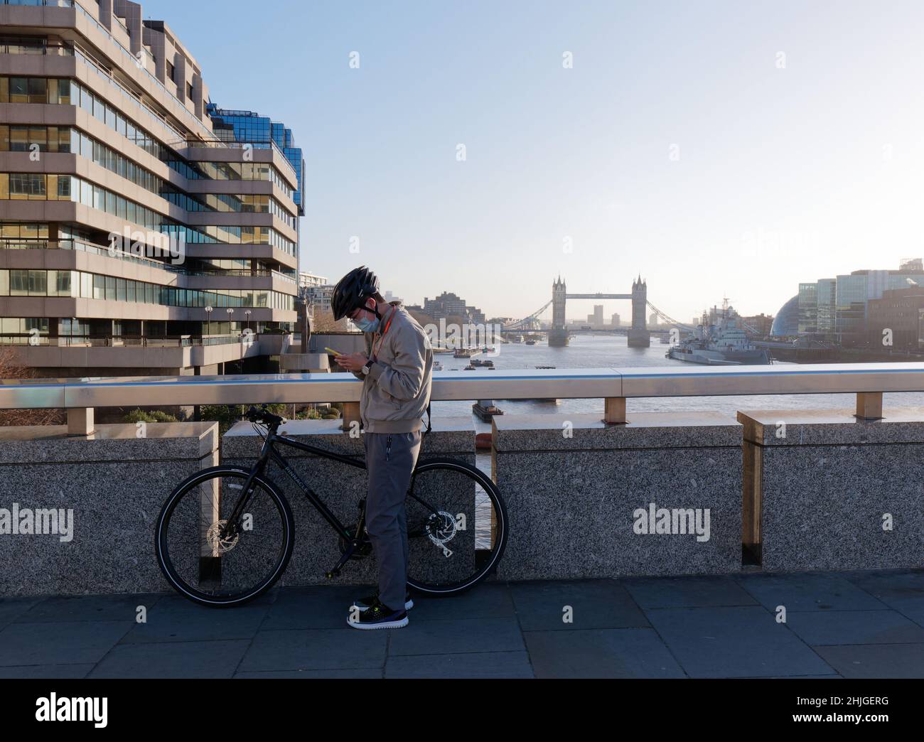 Londres, Grand Londres, Angleterre, 5 janvier 2022: Cycliste portant un masque pendant les contrôles covid téléphone sur le pont de Londres avec Tower Bridge derrière. Banque D'Images