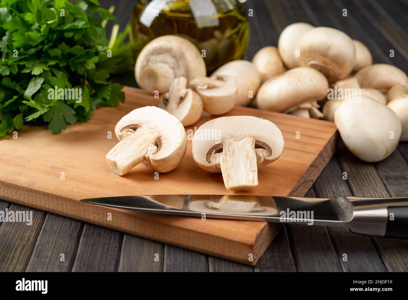 Champignon cru coupé en deux et couteau de cuisine sur une planche à découper, champignons de Paris entiers et bouquet de persil sur fond sombre.Plats végétariens Banque D'Images