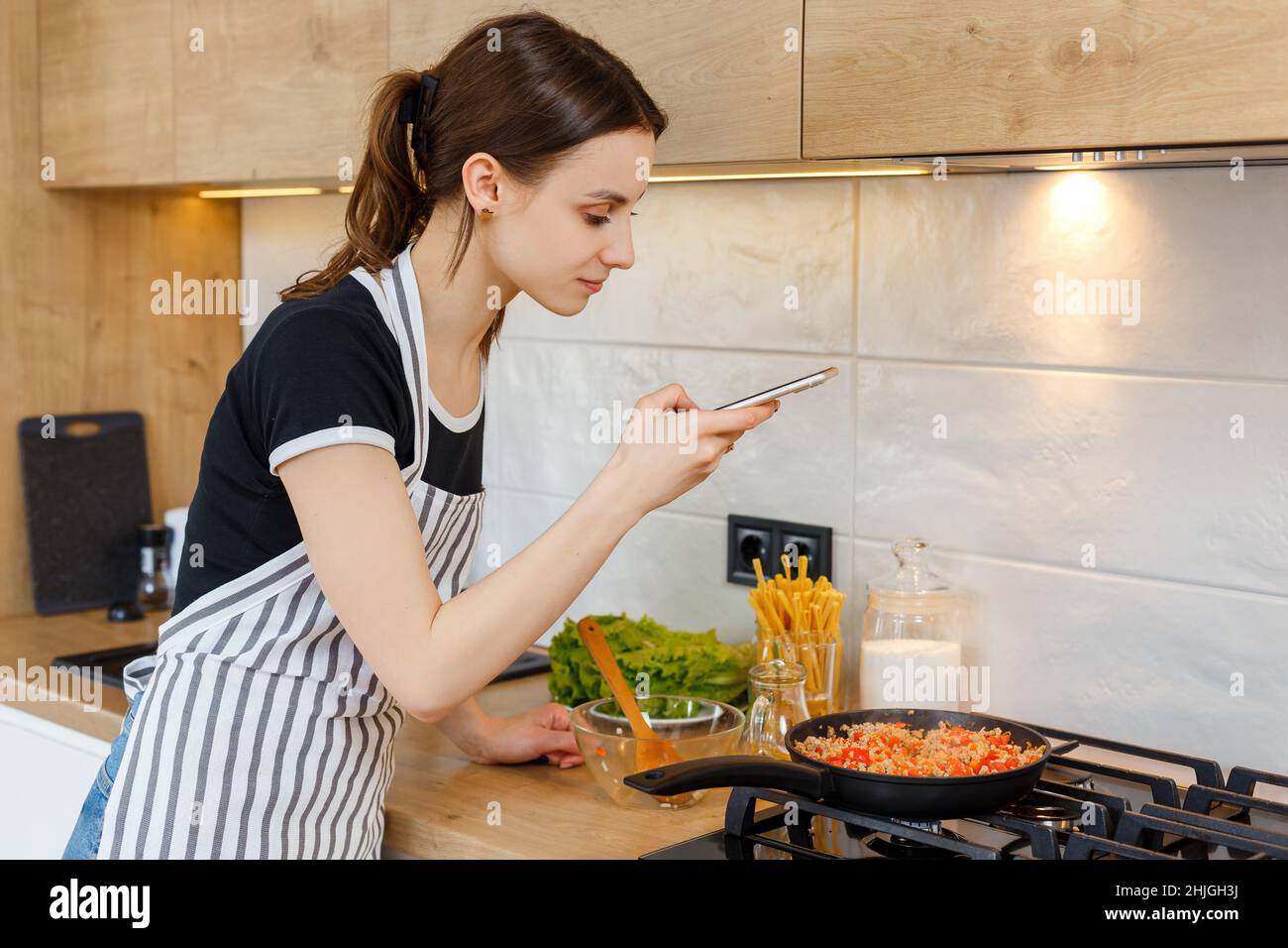 Une jeune femme blogueuse en tablier prend des photos avec le téléphone tout en cuisinant de la nourriture dans la cuisine.Préparation du repas avec une poêle à frire sur une cuisinière à gaz.Concept de style de vie domestique, de loisirs au foyer et de blogging culinaire. Banque D'Images