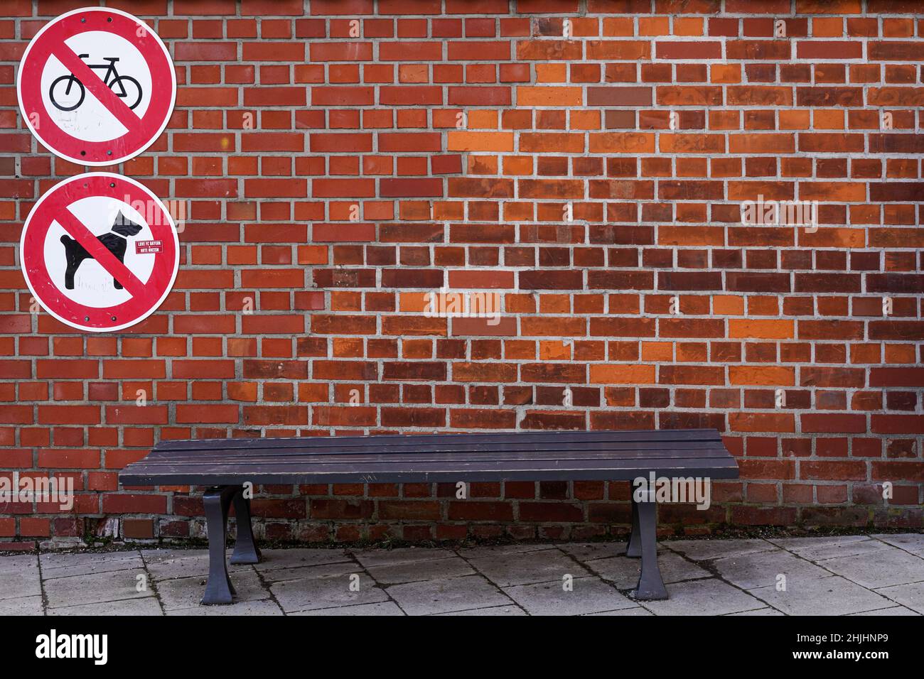 Un banc en bois devant un mur de briques.Des panneaux d'interdiction rouges interdisent le cyclisme et l'introduction de chiens. Banque D'Images