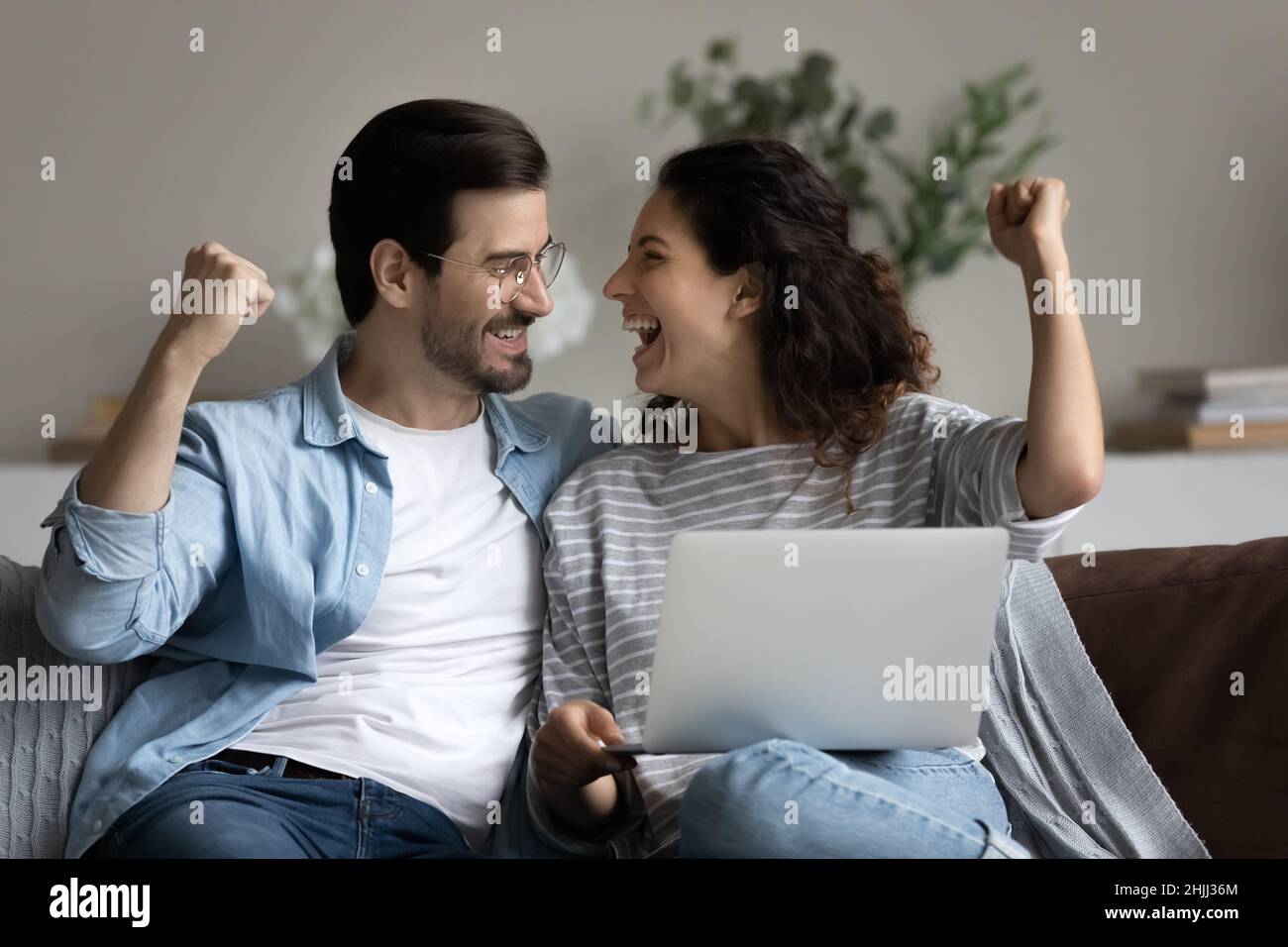 Couple assis sur un canapé avec un ordinateur portable, les poings serrés, célébrer le succès Banque D'Images