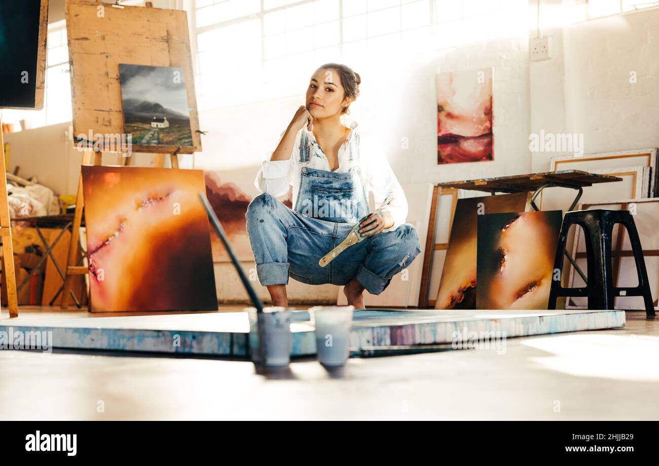 Une jeune artiste s'accroupir sur le sol tout en travaillant dans son studio d'art.Femme peintre regardant la caméra et tenant un pinceau.Jeune wom créatif Banque D'Images