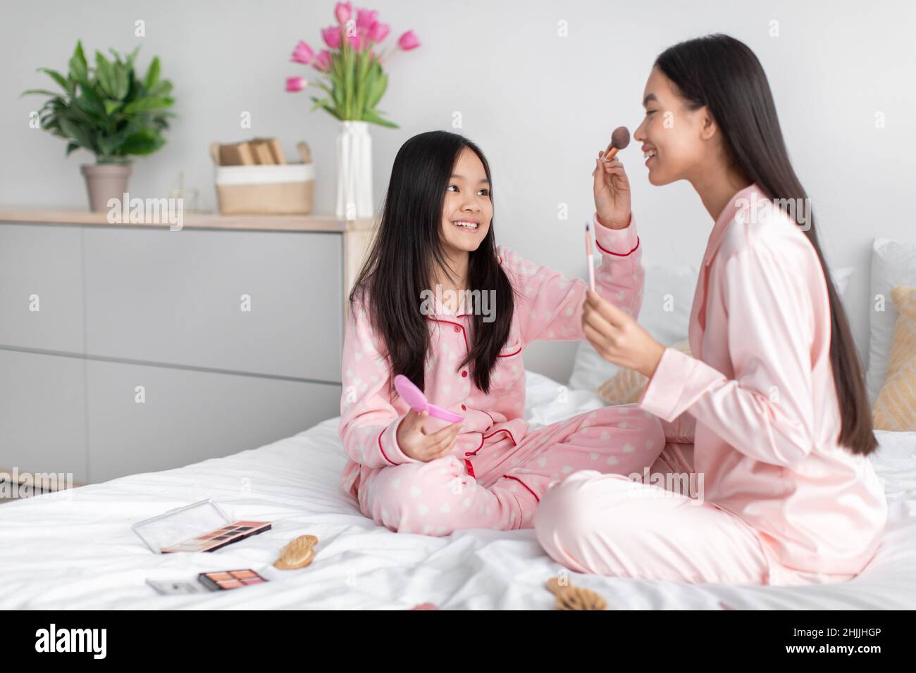 Joyeuse et heureuse jeune fille coréenne appliquant de la poudre au visage de femme millénaire, assise sur le lit avec des cosmétiques Banque D'Images