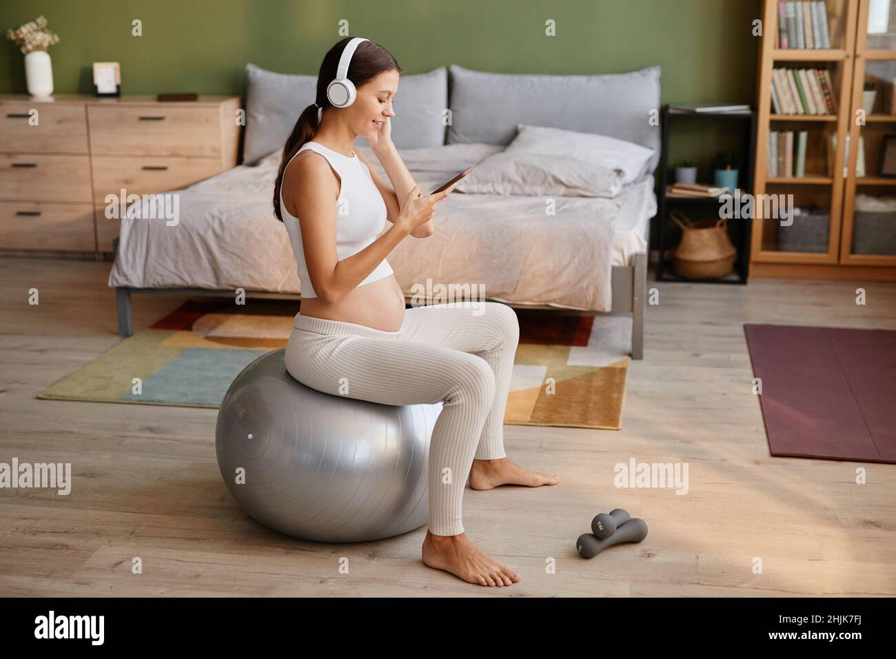 Jeune femme enceinte dans un casque utilisant un téléphone portable pour écouter de la musique tout en faisant de l'exercice sur le ballon de fitness dans la chambre Banque D'Images
