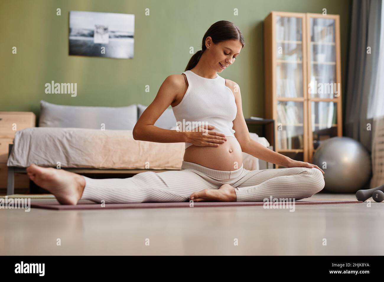 Heureuse femme enceinte assise sur le tapis d'exercice et touchant son ventre, elle se préparant à l'accouchement faisant des exercices d'étirement dans la chambre Banque D'Images