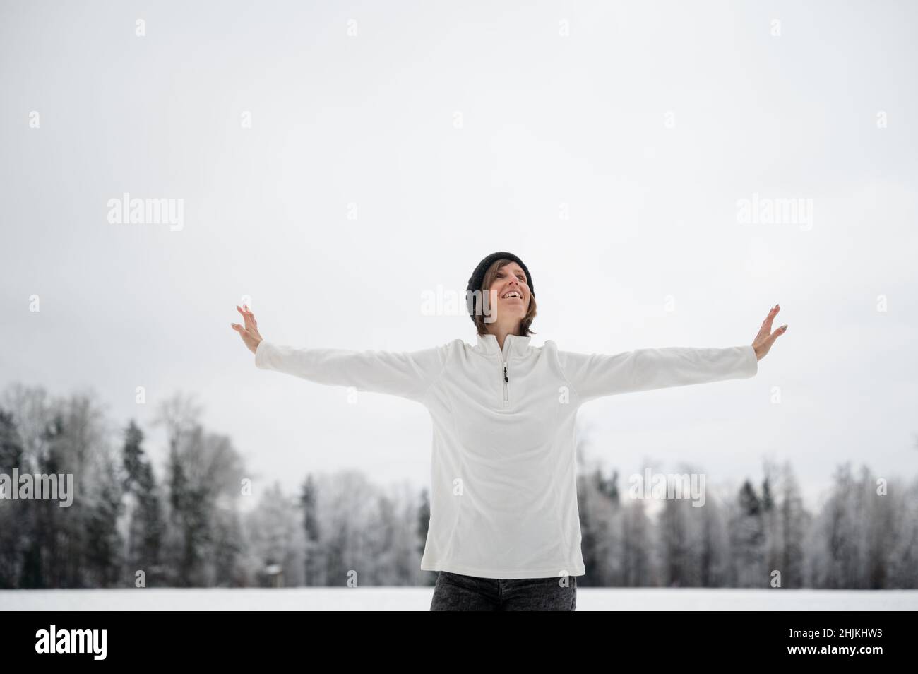 Jeune femme souriante en chandail blanc debout avec ses bras se propageant au milieu de la nature enneigée de l'hiver, célébrant la vie. Banque D'Images