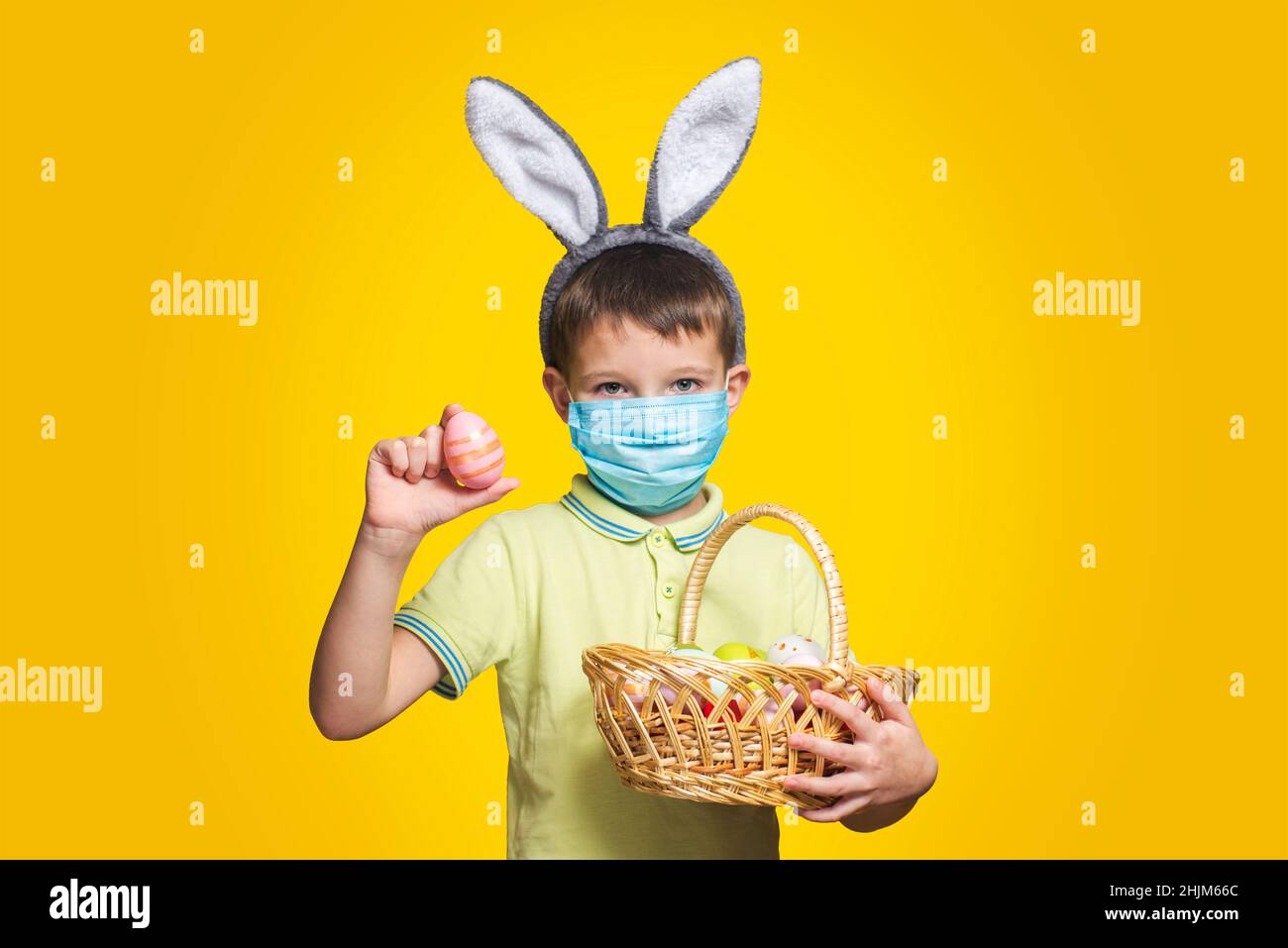 Petit garçon portant un masque médical jetable avec des oreilles de lapin, tenant un panier d'oeufs de Pâques sur un fond jaune.Jour de Pâques pendant le coronavirus Banque D'Images