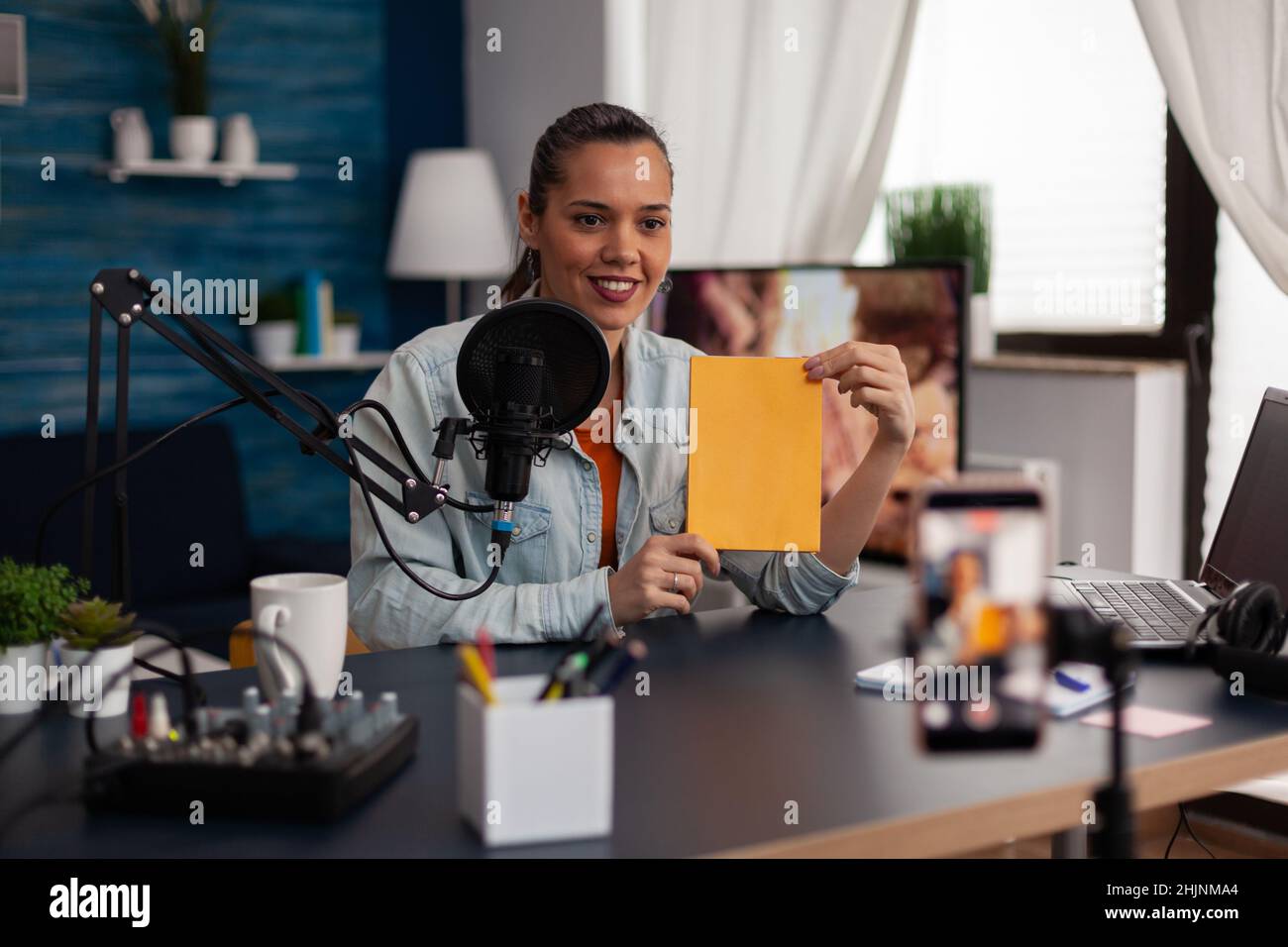 Femme influenceur présentant un livre jaune devant un smartphone d'enregistrement.Similing content Creator diffusant un commentaire sur les réseaux sociaux dans un studio à domicile avec microphone et ordinateur portable. Banque D'Images