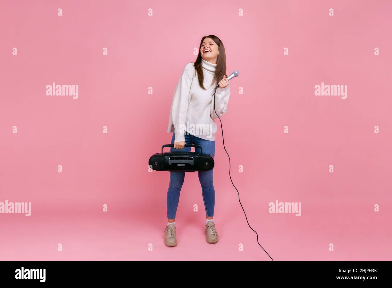 Portrait complet de la femme heureuse optimiste tenant un lecteur de disques et un microphone, chantant des chansons, portant un chandail blanc de style décontracté.Studio d'intérieur isolé sur fond rose. Banque D'Images