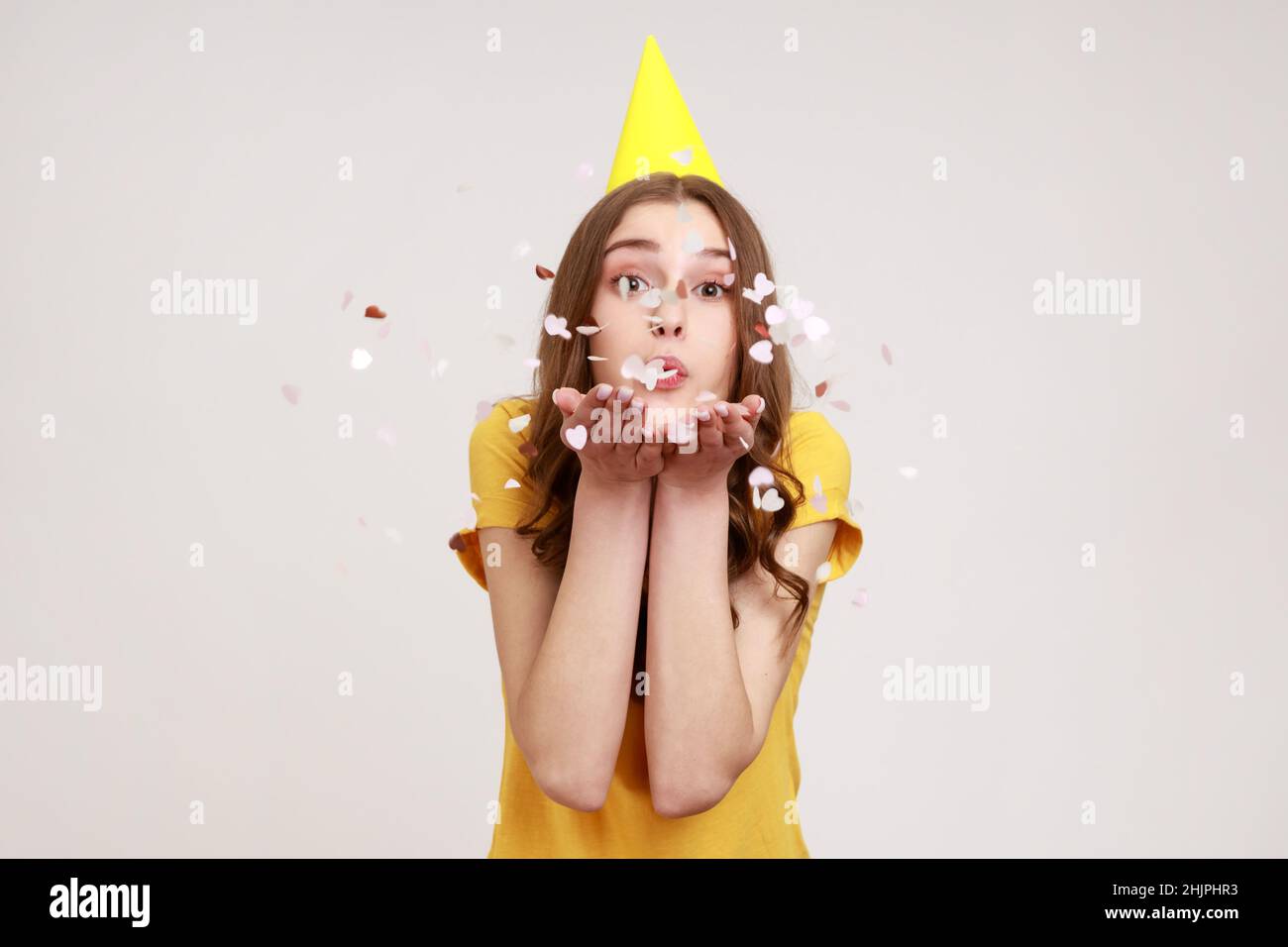 Insouciante adolescente heureuse fille dans un t-shirt jaune décontracté et chapeau de fête scintillant, profiter de l'anniversaire, célébrer l'événement, ambiance festive.Prise de vue en studio isolée sur fond gris. Banque D'Images