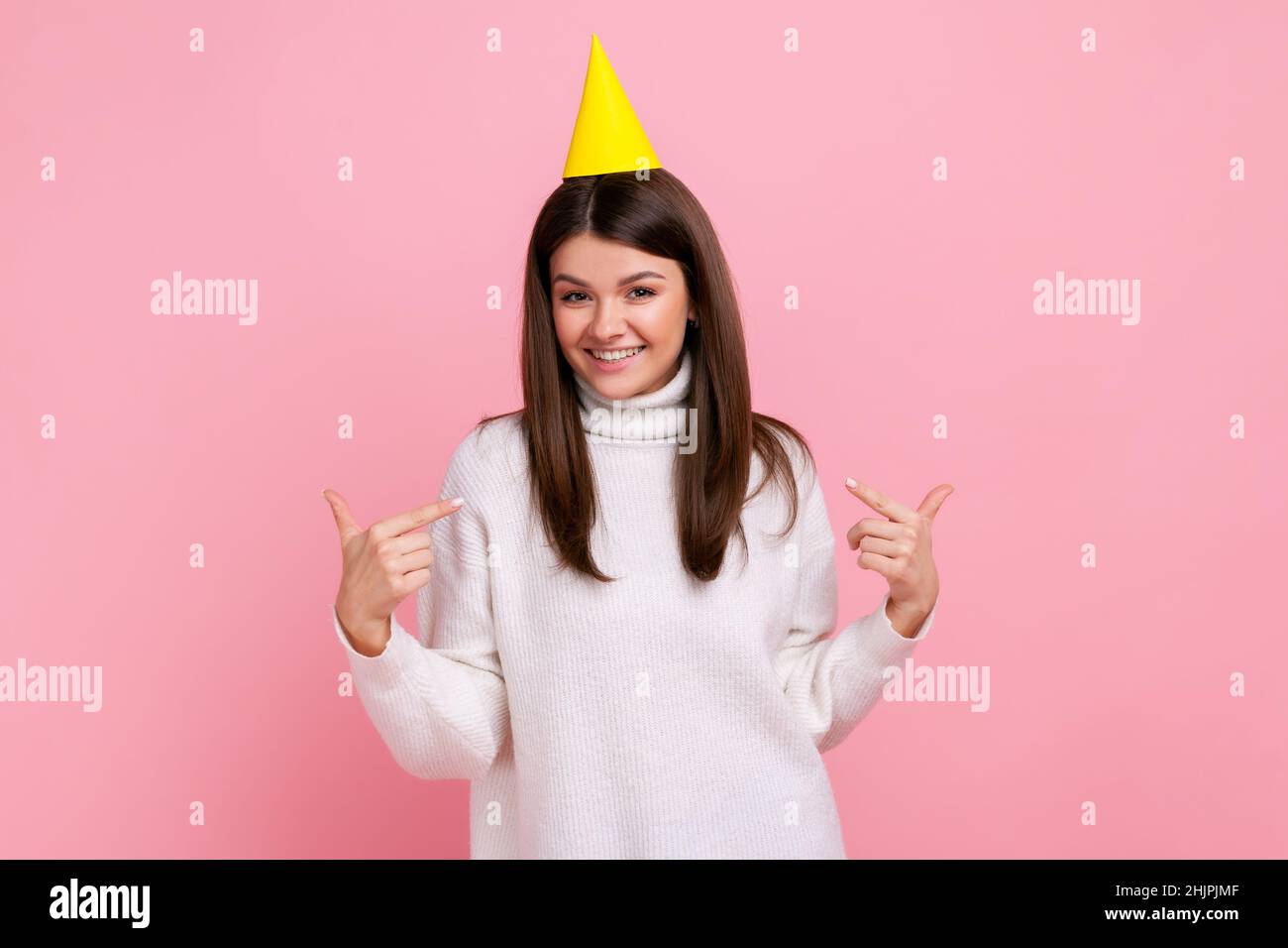 Portrait d'une fille brunette heureuse et satisfaite dans un cône de fête se pointant vers elle-même, célébrant son anniversaire, portant un chandail blanc de style décontracté.Studio d'intérieur isolé sur fond rose. Banque D'Images