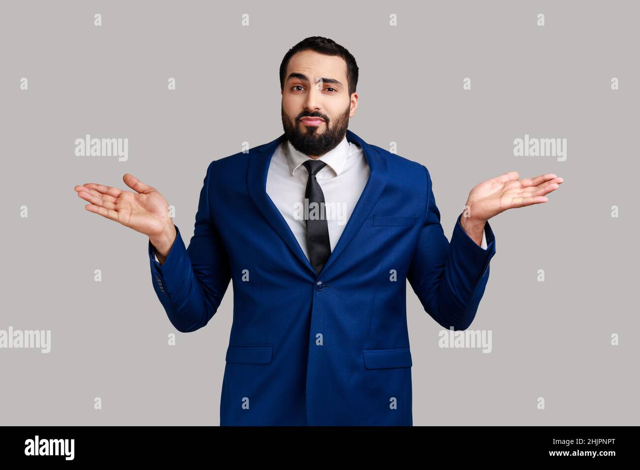 Portrait d'un jeune homme d'affaires barbu adulte confus haussant les épaules, ne faisant aucun geste d'idée, quoi qu'il en soit, portant un costume de style officiel.Prise de vue en studio isolée sur fond gris. Banque D'Images