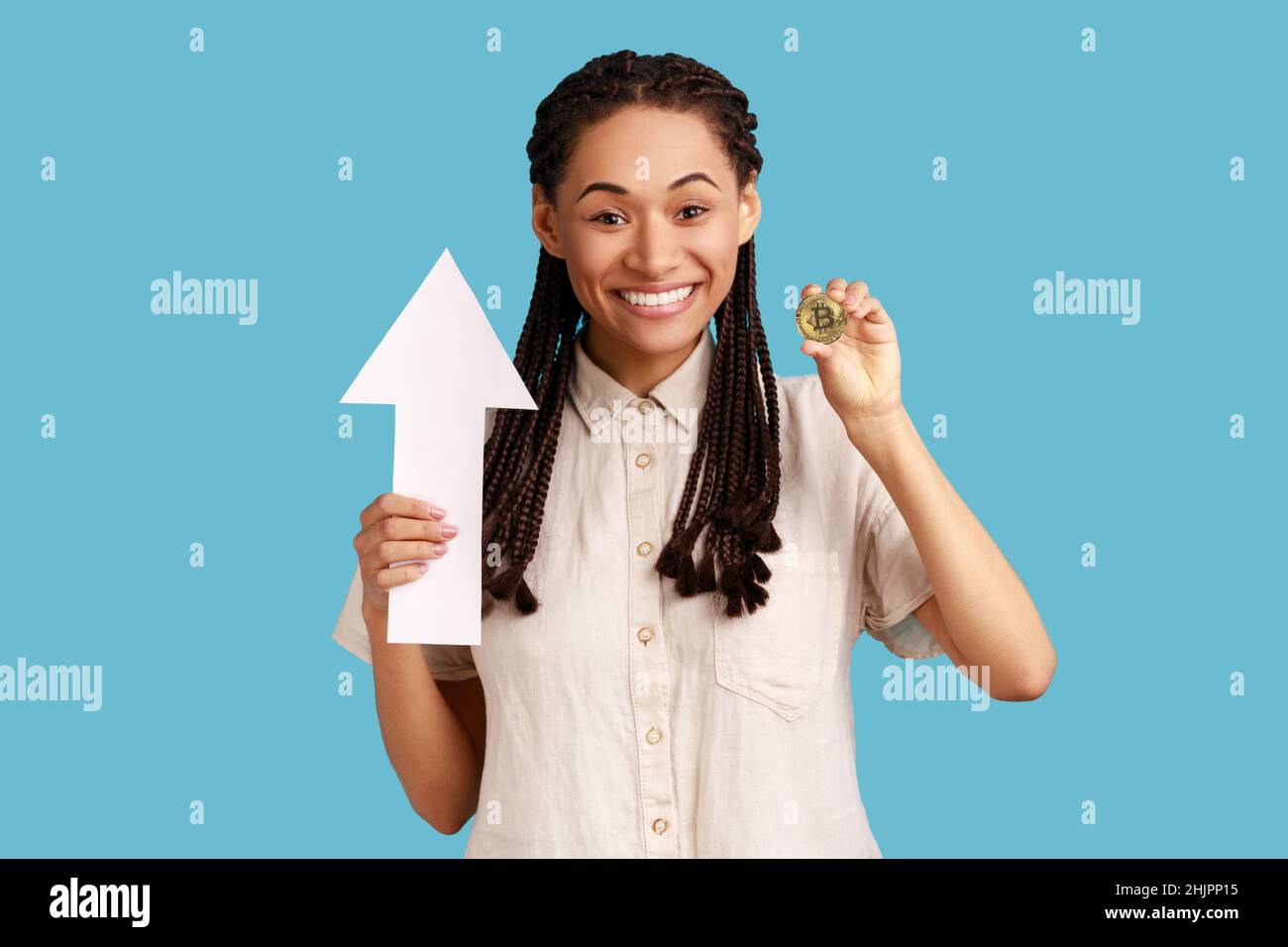 Portrait d'une femme satisfaite avec des dreadlocks noirs montrant le bitcoin d'or et la flèche pointant vers le haut, la croissance de la monnaie électronique, portant une chemise blanche.Studio d'intérieur isolé sur fond bleu. Banque D'Images