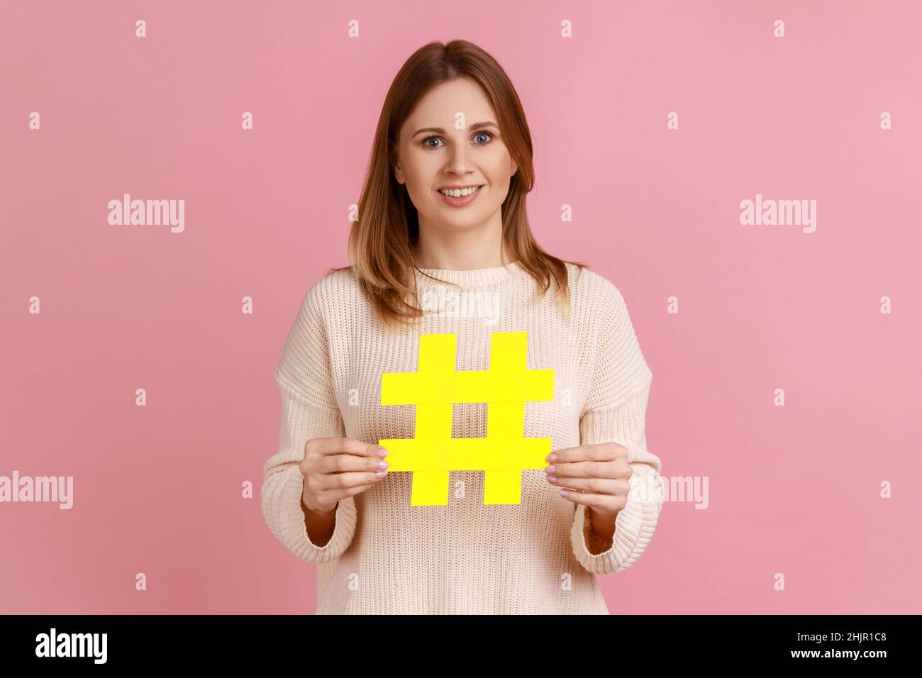 Portrait d'une femme blonde optimiste tenant un grand symbole jaune hashtag et souriant à la caméra, promotion des tendances du blog, portant un chandail blanc.Studio d'intérieur isolé sur fond rose. Banque D'Images