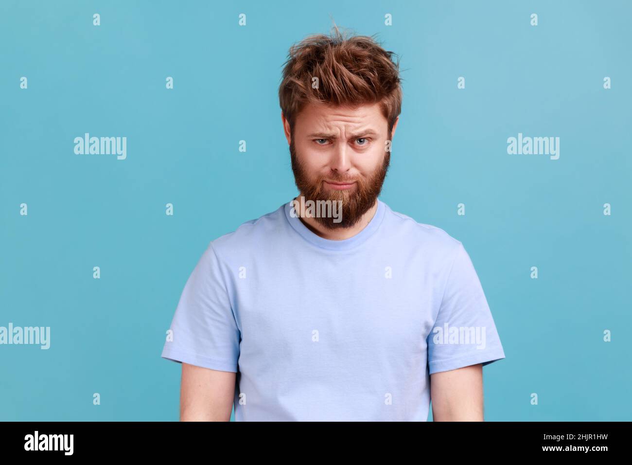 Portrait du jeune homme en colère barbu debout et regardant la caméra avec le visage de tristesse insatisfait, exprimant la douleur, ayant mauvaise humeur.Studio d'intérieur isolé sur fond bleu. Banque D'Images