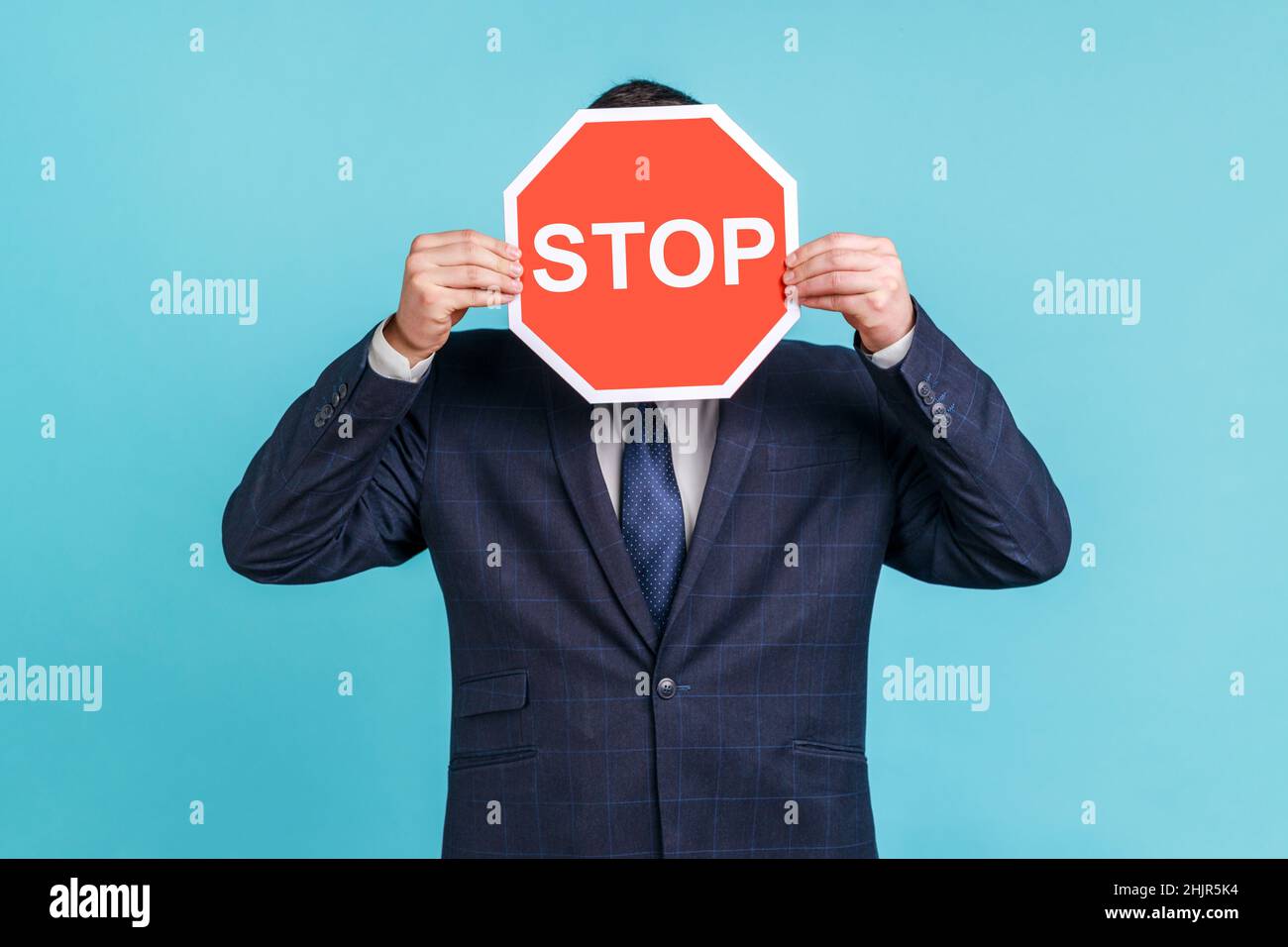 Portrait d'un employé de bureau inconnu portant un costume officiel cachant son visage derrière un panneau d'arrêt rouge, avertissement de danger, restriction et limites.Studio d'intérieur isolé sur fond bleu. Banque D'Images